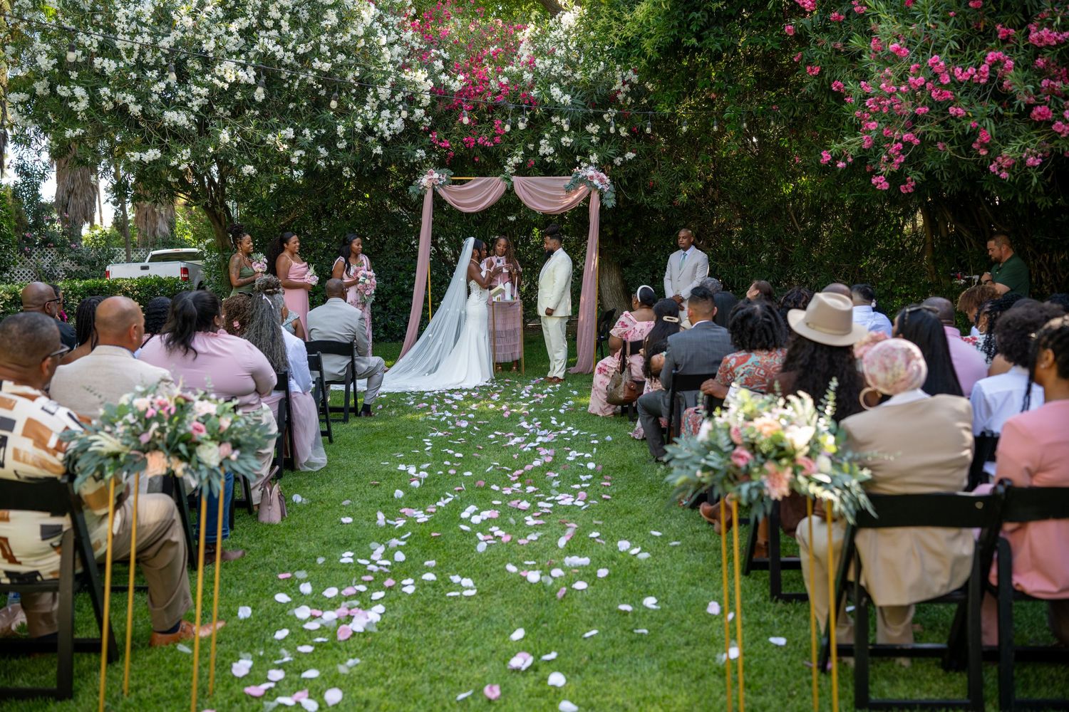 An outdoor wedding with guests seated on chairs along a floral-lined aisle.
