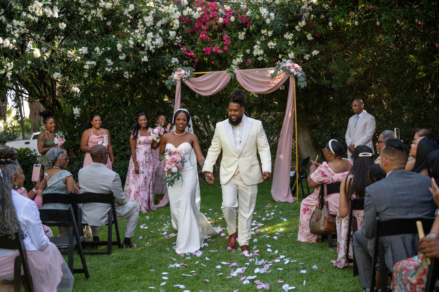 Wedding guests gather in a garden setting with scattered rose petals on the ground.