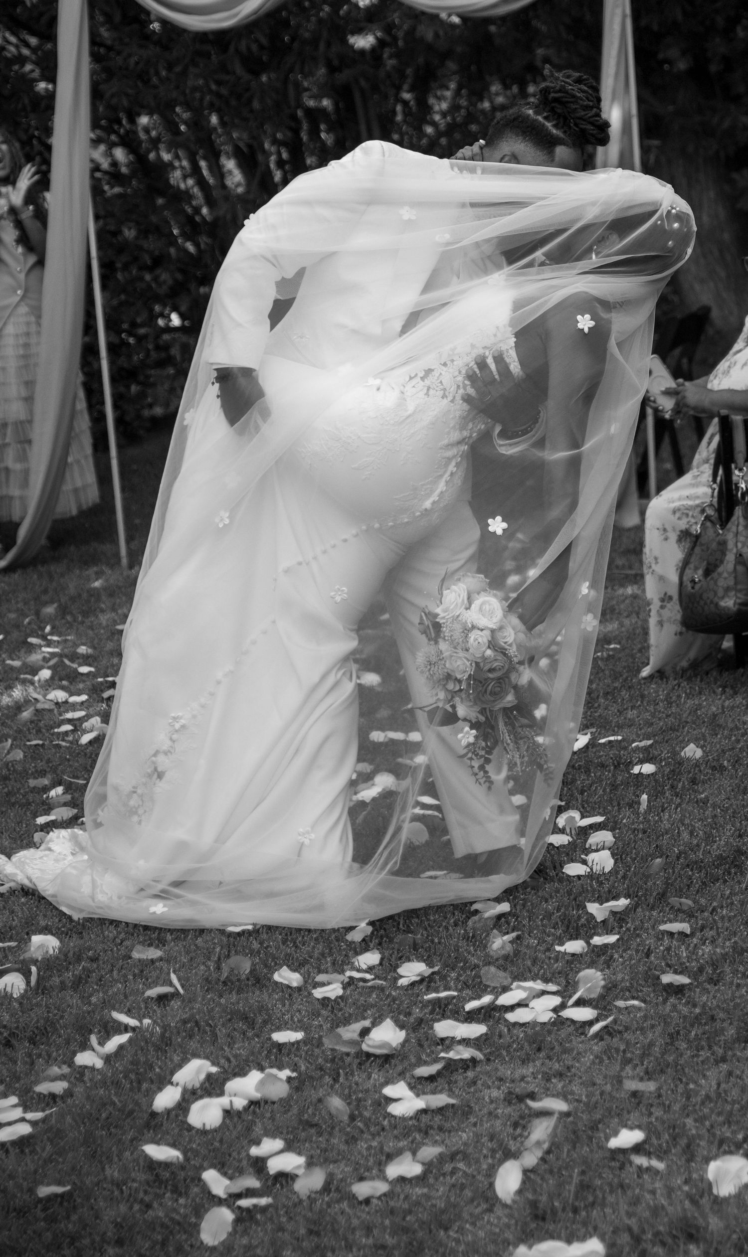A flowing white wedding dress and veil trail on grass covered with scattered petals.
