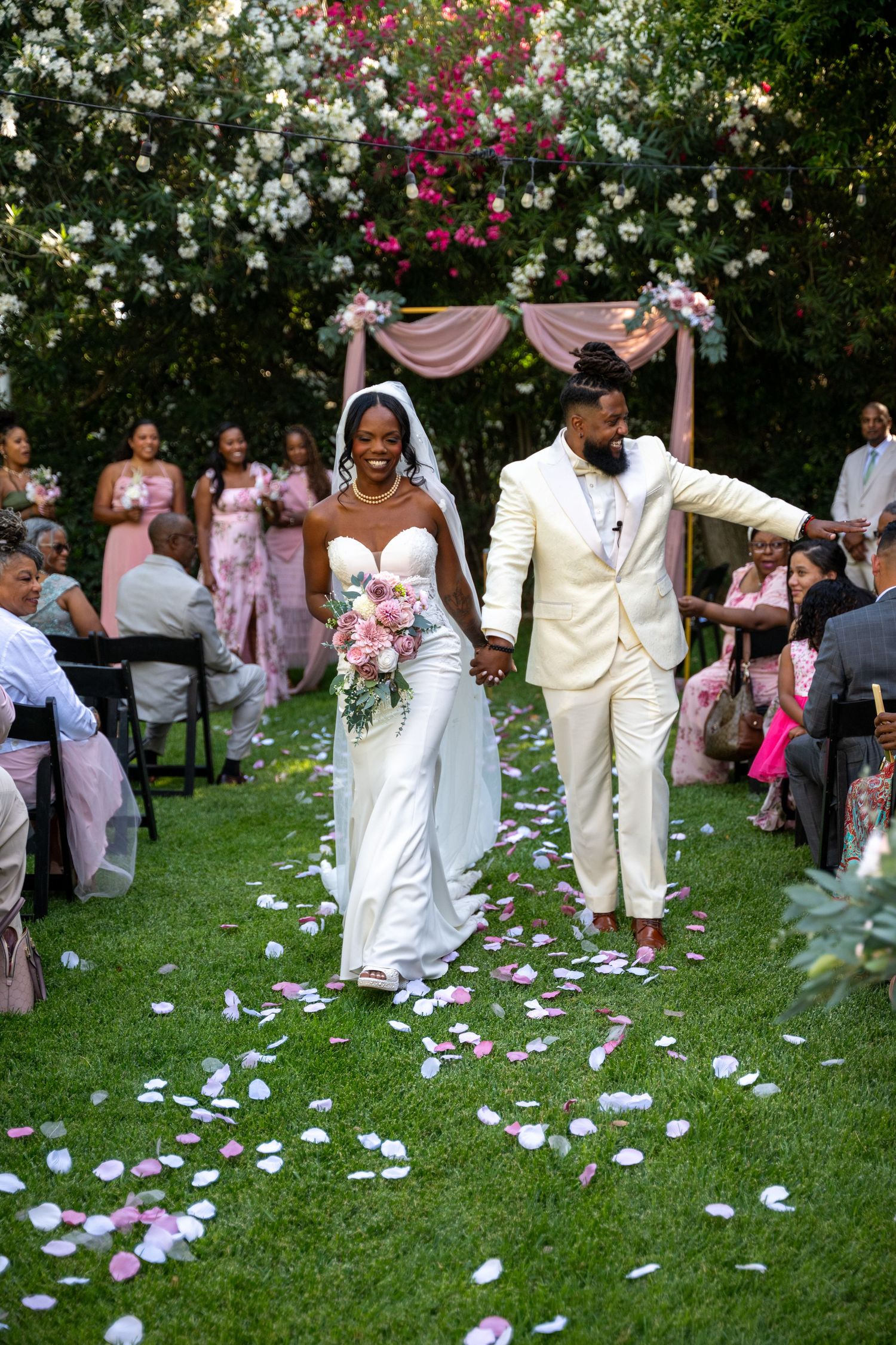 Wedding party members in white and cream attire walk through a garden setting.