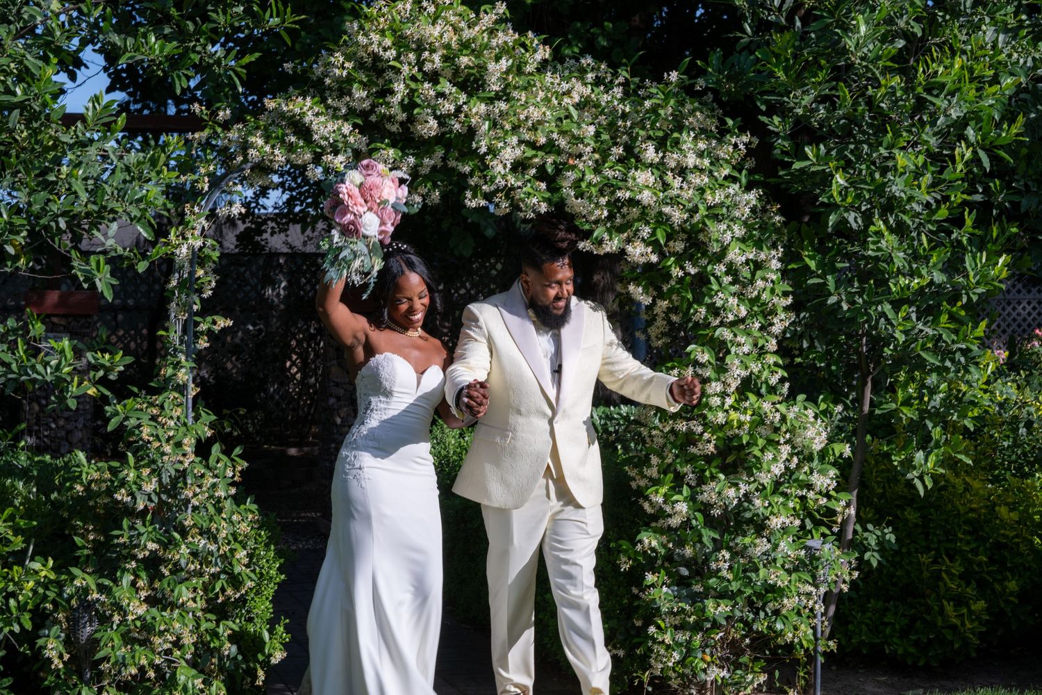 A wedding couple celebrates joyfully in front of a floral garden arch on their special day.