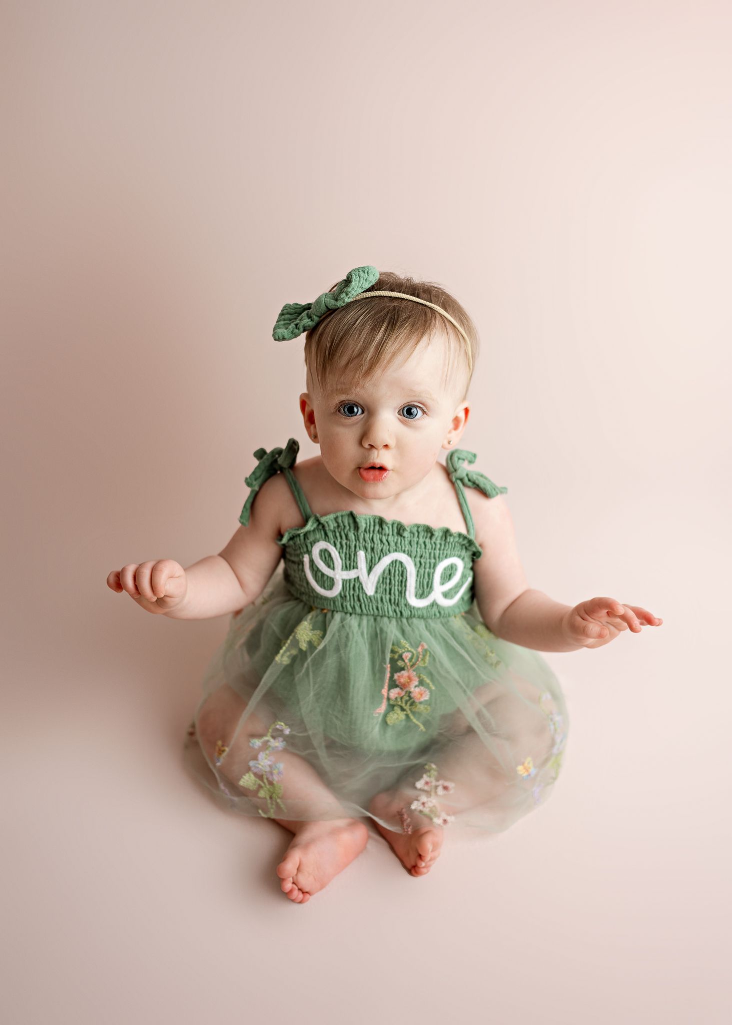 Baby wearing a mint green floral dress with 'one' text and green bow accessories sits on beige backdrop for first birthday photo.