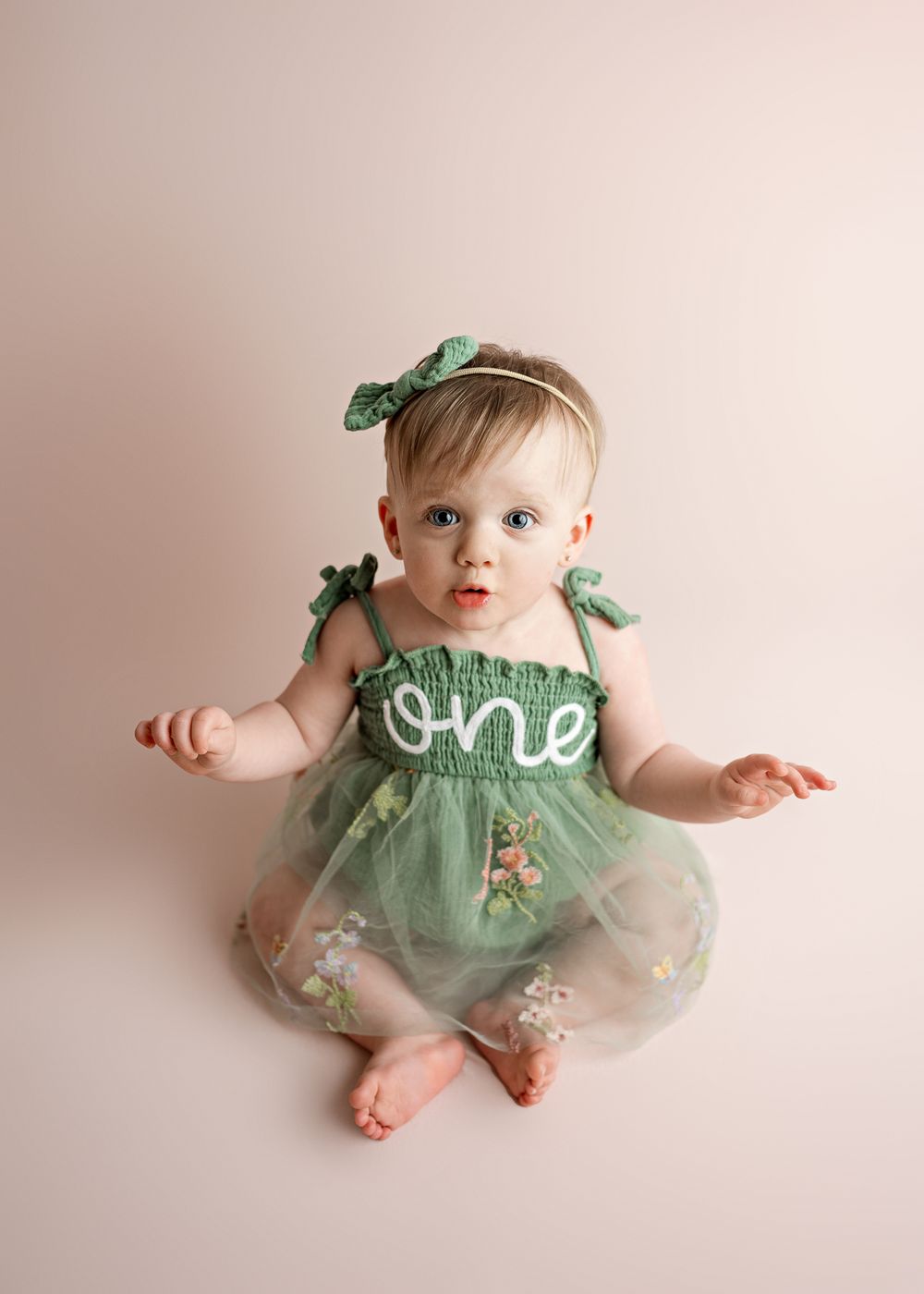 Baby wearing a mint green floral dress with 'one' text and green bow accessories sits on beige backdrop for first birthday photo.