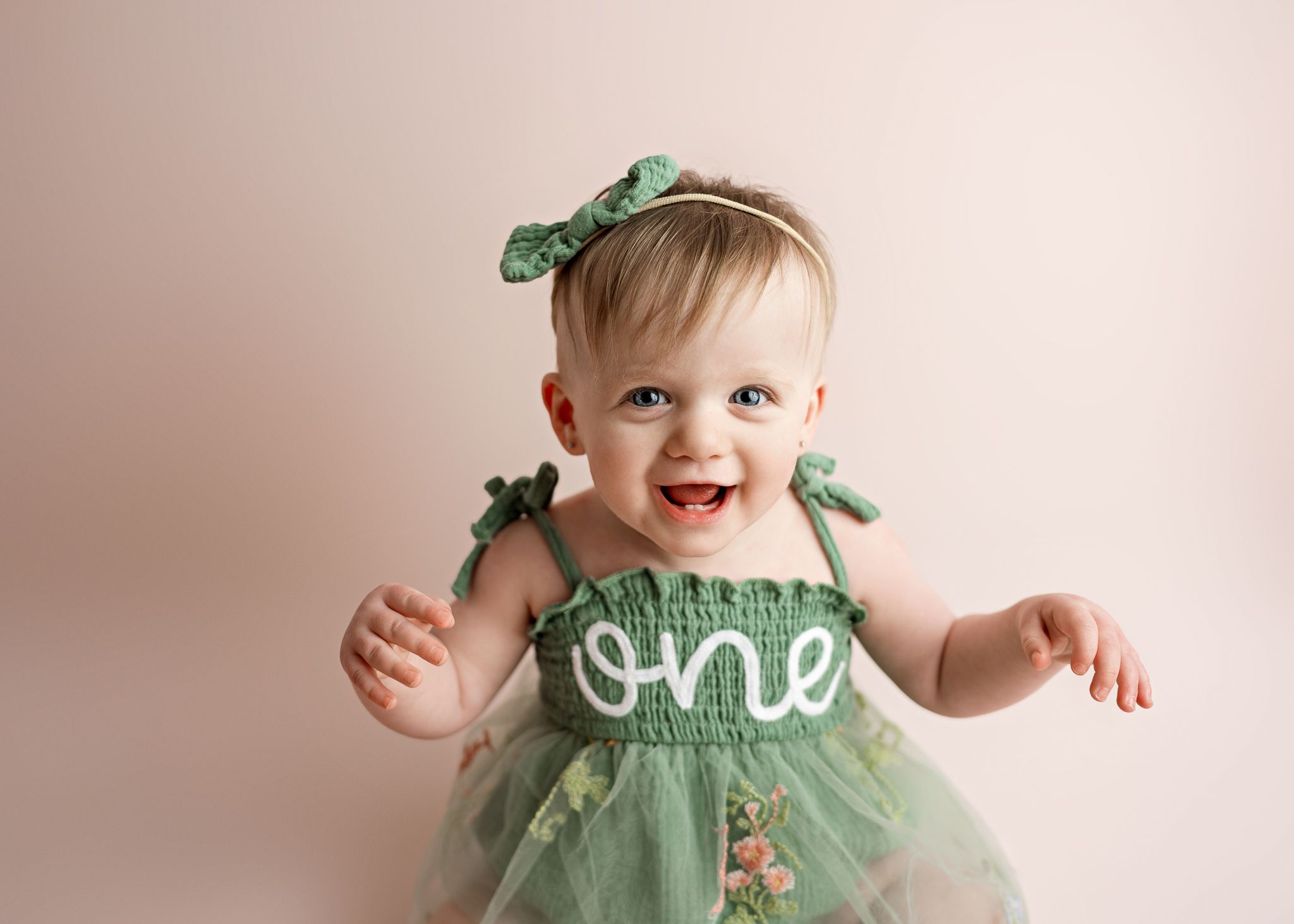Happy baby in mint green tulle dress with the word 'one' wearing matching hair bows against beige background.