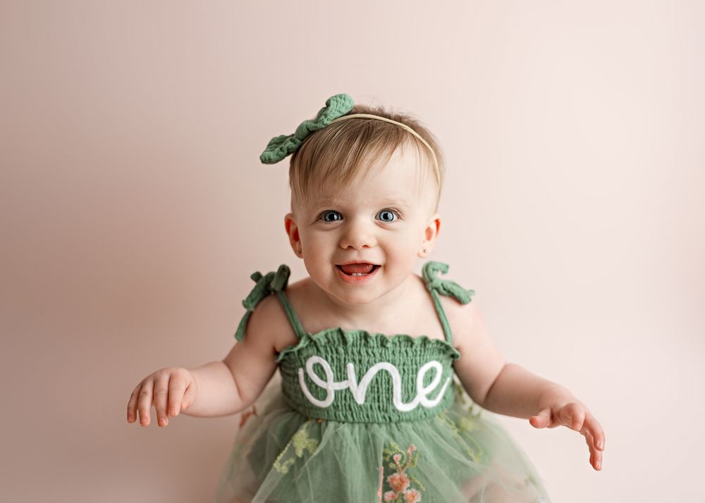 Small baby wearing a mint green dress with bows and \"one\" text detail smiles against beige backdrop.