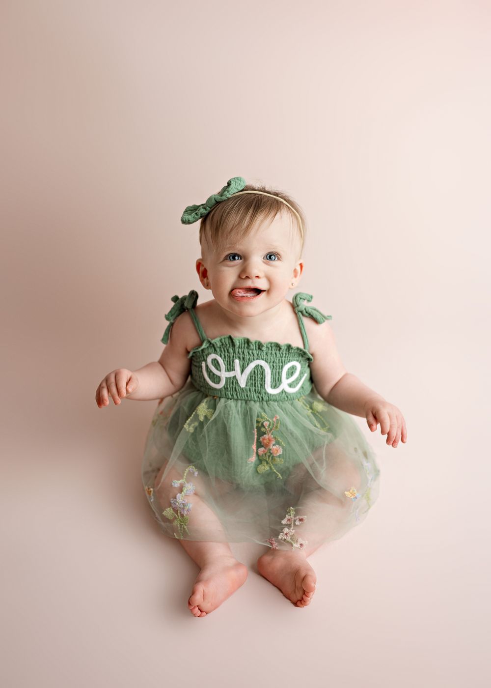 Baby in mint green tulle dress with 'one' text and matching bow sitting on beige background.