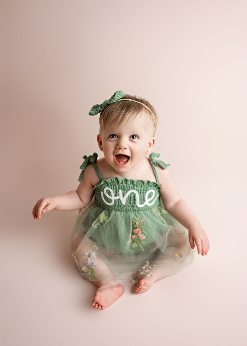Small child in mint green tulle dress with 'one' text design crawls and poses against light pink backdrop during photoshoot.
