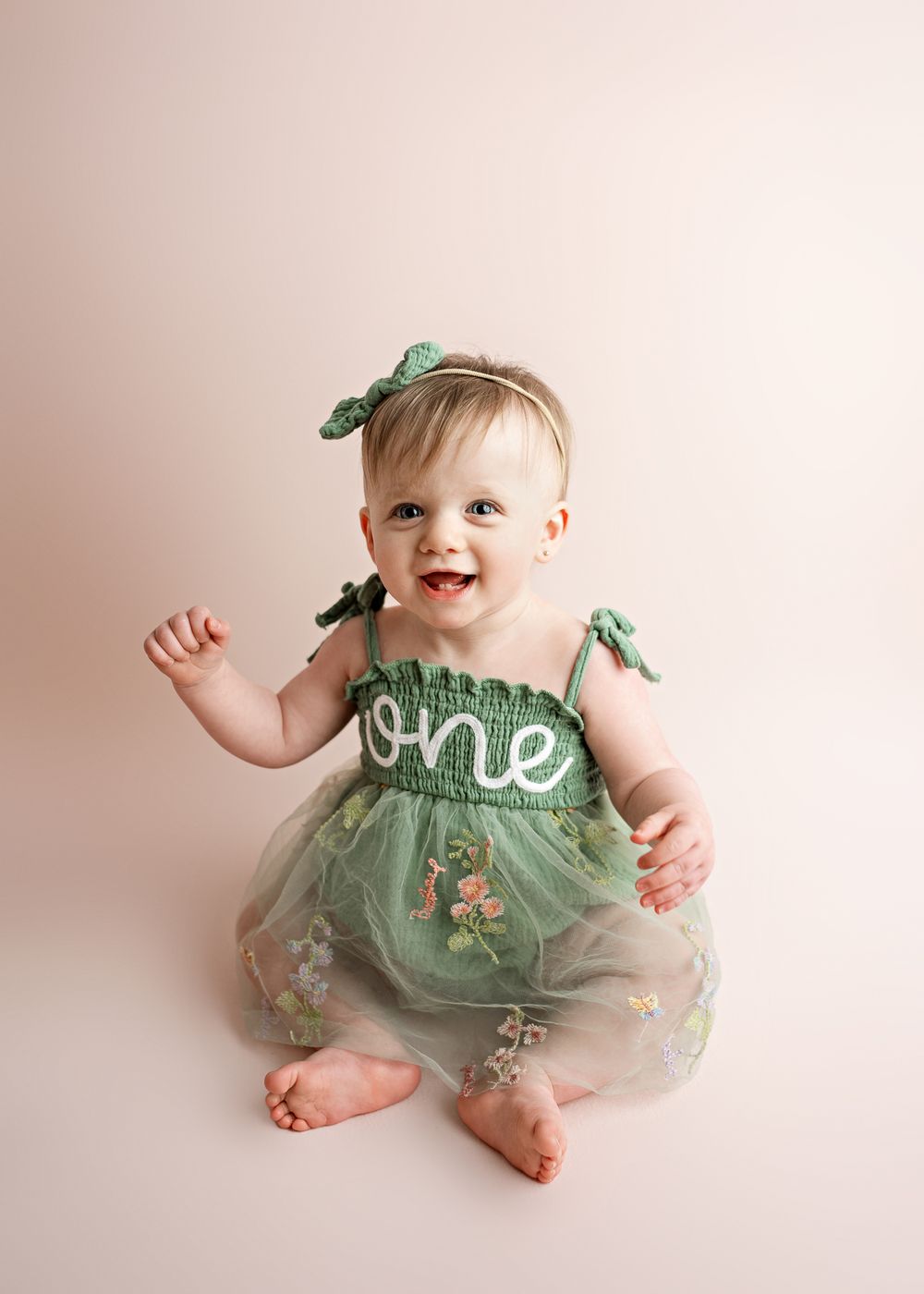 Baby in a sage green dress with floral details smiling and sitting against a soft beige background.