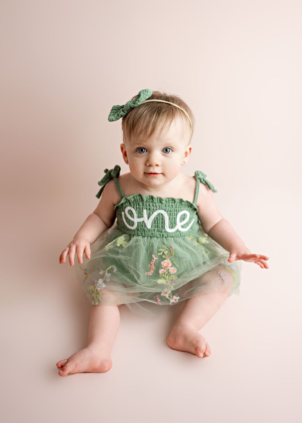 Baby wearing mint green tulle dress with 'one' text and matching bow headband sits against pink background.