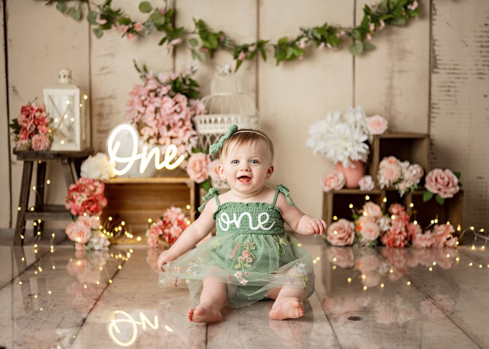 A festive first birthday setup with mint dress, floral decorations, fairy lights and a neon 'One' sign against rustic backdrop.