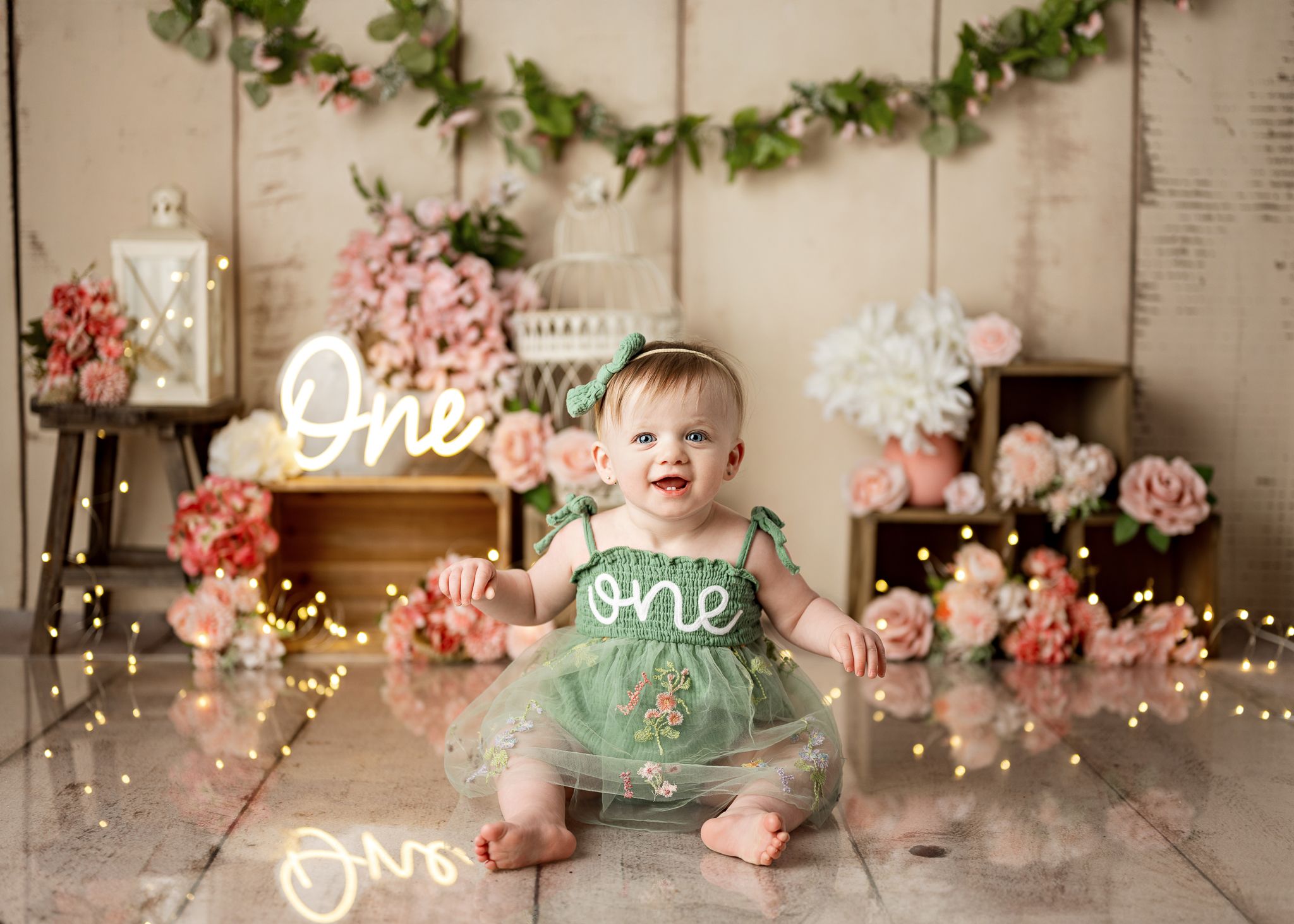 First birthday photo setup with mint green outfit, floral decorations, string lights and neon 'one' sign against wooden backdrop.