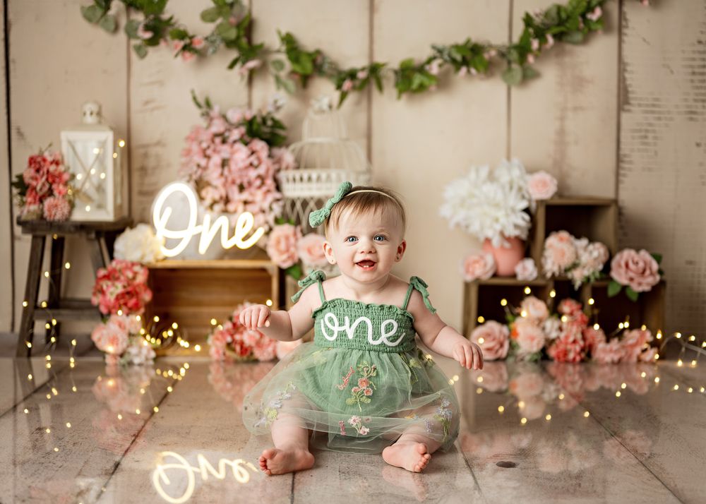 First birthday photo setup with mint green outfit, floral decorations, string lights and neon 'one' sign against wooden backdrop.