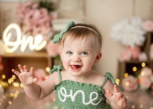 A baby in green attire sits smiling with a glowing 'One' sign and pink flowers in the background.