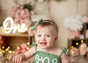 Baby in green outfit smiles against pink floral backdrop with glowing 'One' neon sign and bokeh lights.