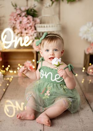 A baby in a mint green tulle dress sits on a carpeted floor with string lights and a neon 'One' sign in the background.