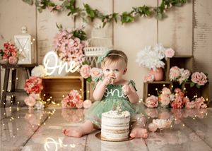 A first birthday cake smash photoshoot setup with floral decorations, neon signs, and a mint green outfit.