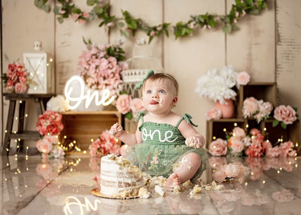 First birthday party setup with rustic decor featuring a small cake, neon signs, floral garlands and pink flowers.