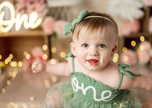 A small child wearing a festive green dress sits in front of twinkling holiday lights and pink decorations.