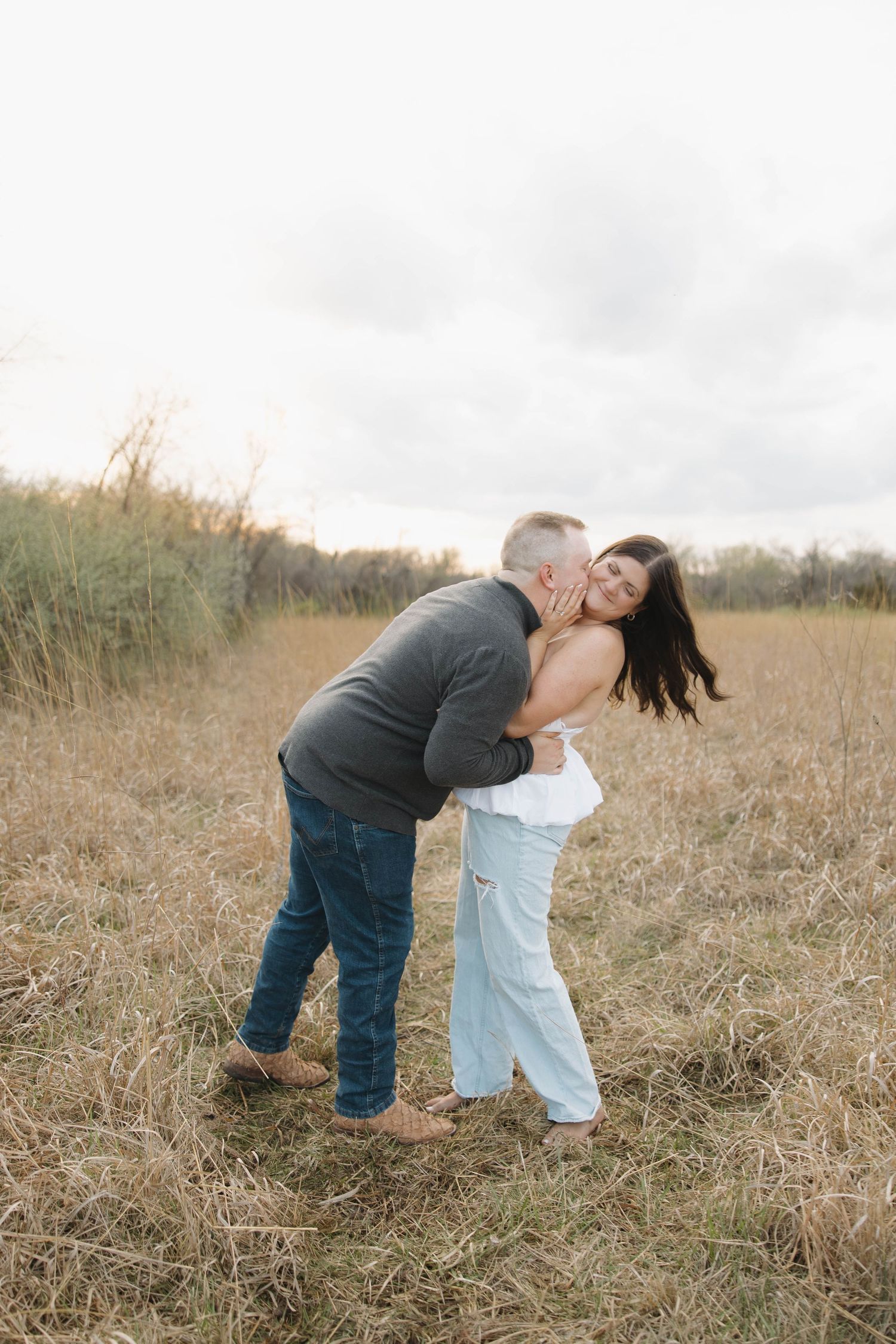 A romantic couple embraces and laughs together in a golden field of tall grass at sunset.