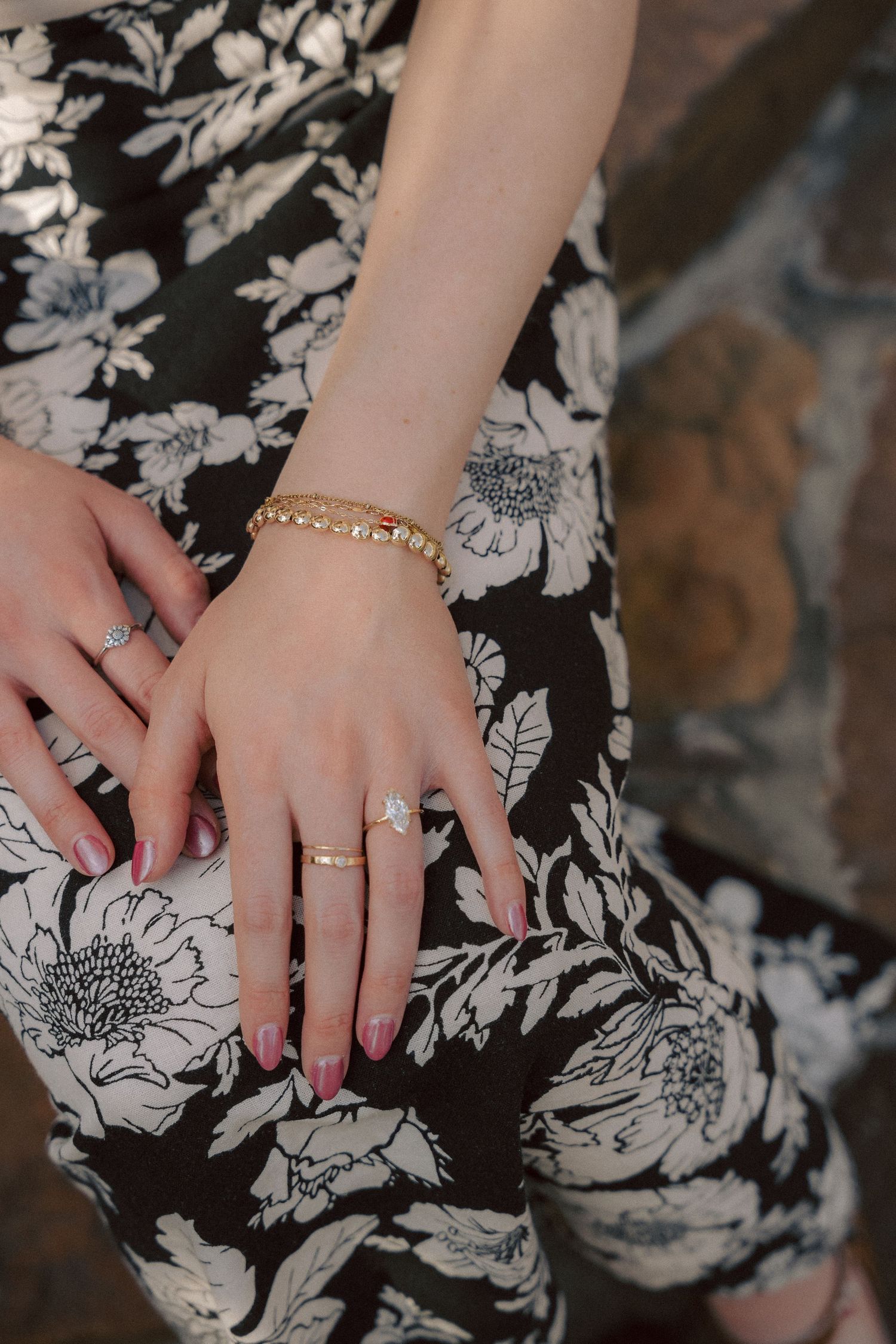 Hands wearing delicate gold jewelry rest on a black and white floral print dress, showing off rings and a bracelet.