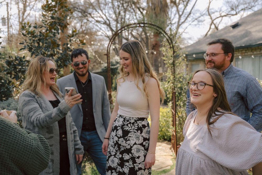 A group of people laughing together outdoors at a casual gathering in a backyard garden setting.