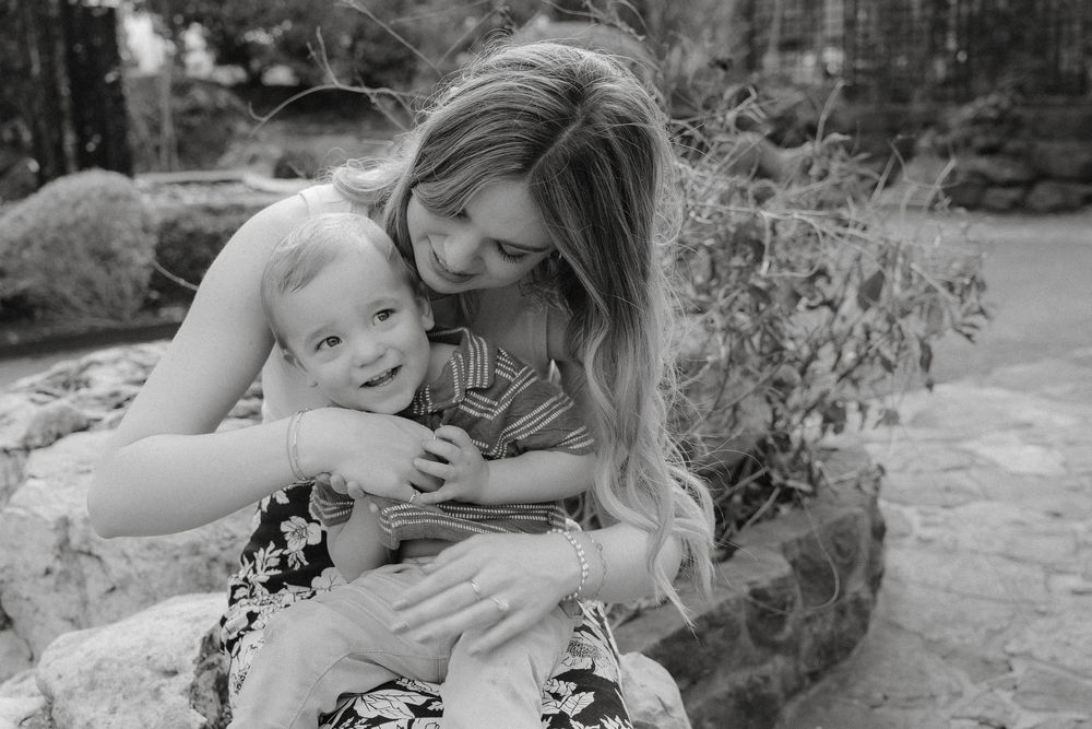 A tender black and white photograph capturing a mother and baby sharing an intimate loving moment outdoors.