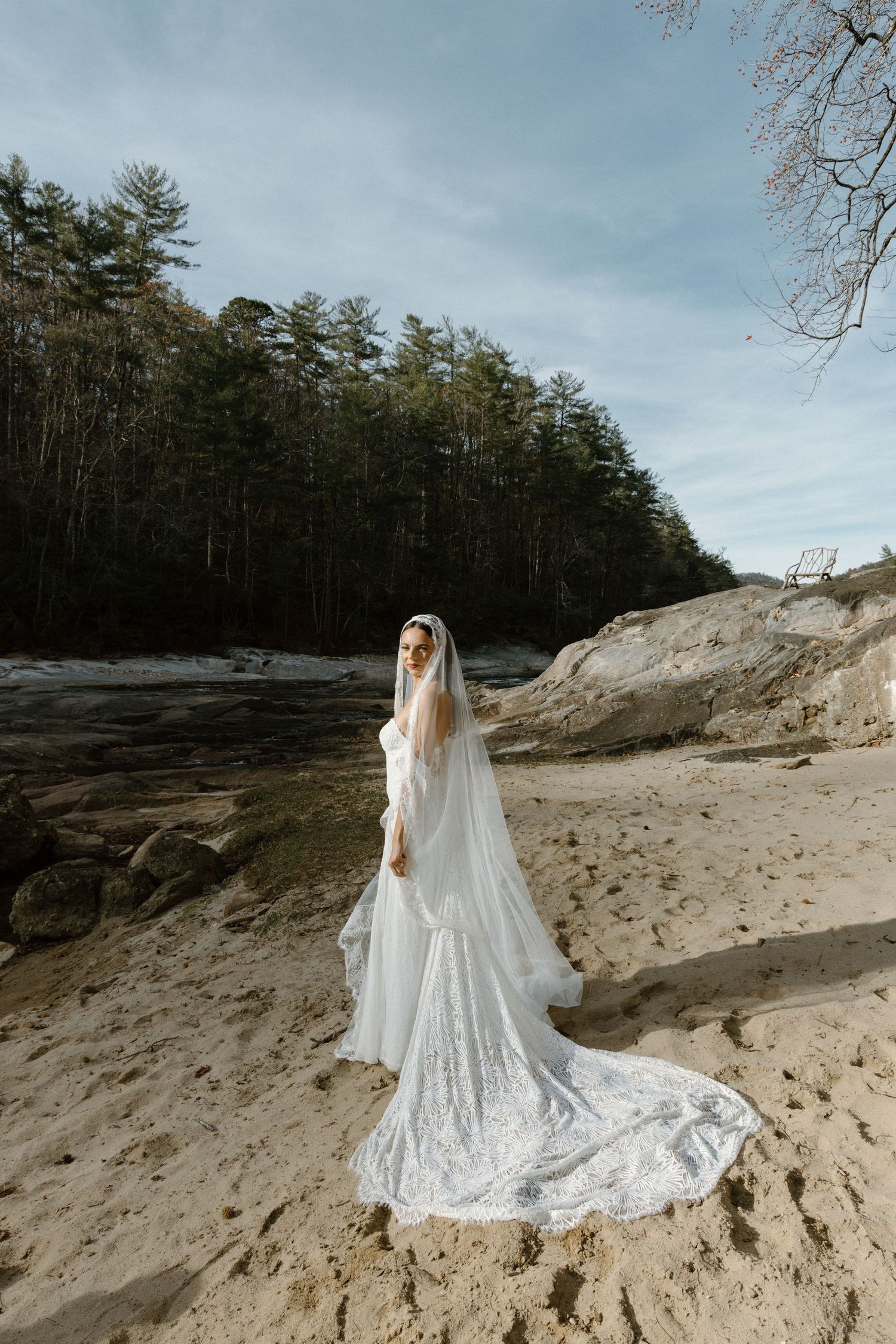 Person in flowing white wedding dress with long train standing on sandy beach near forest.