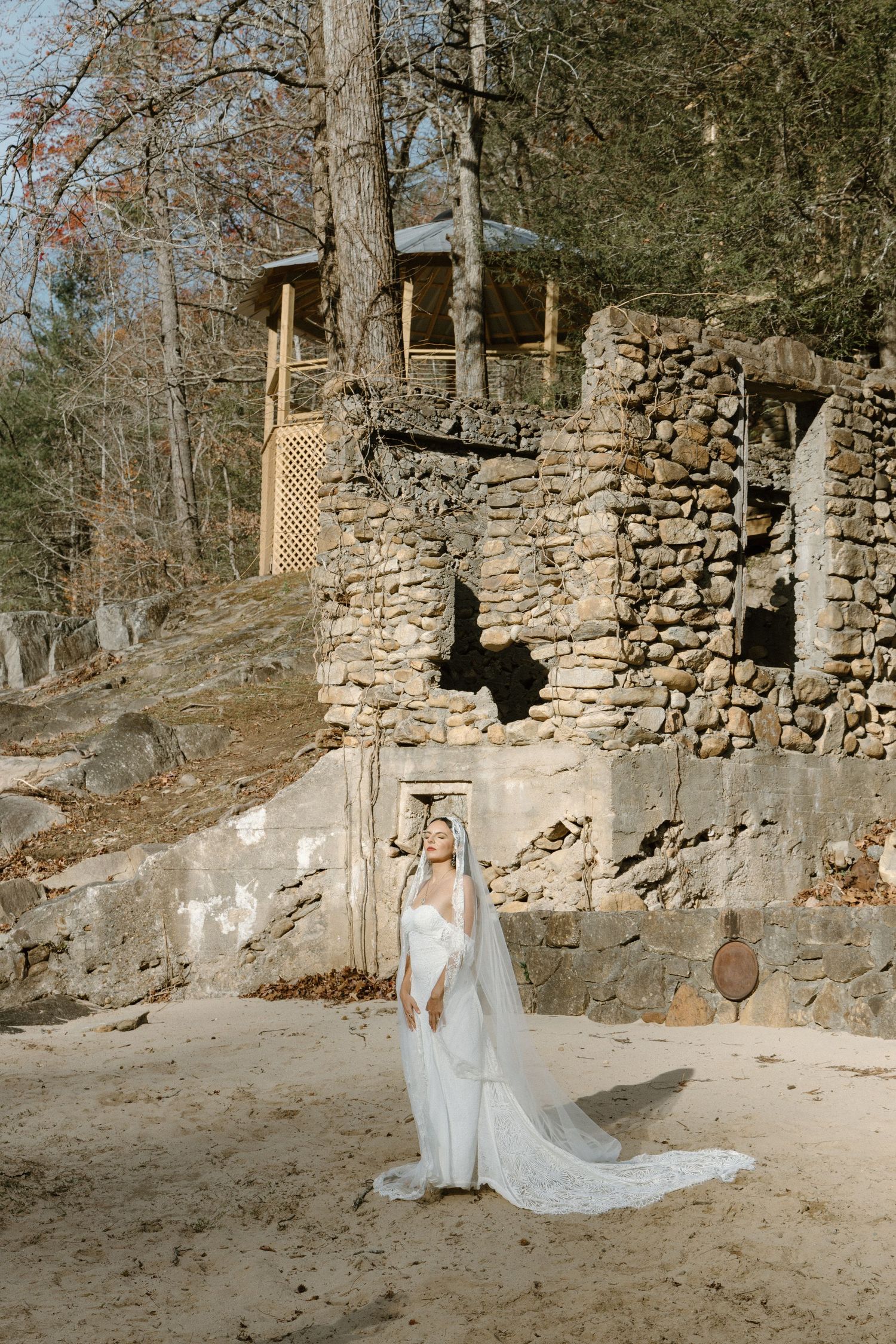 A flowing white wedding dress and veil against a rustic stone wall and woodland cabin backdrop in warm natural light.