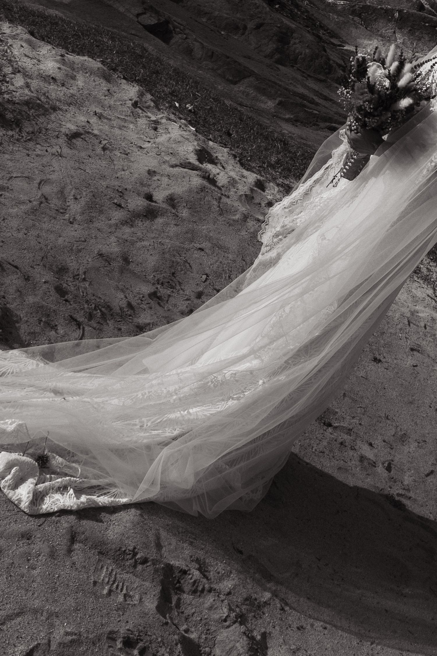 A long flowing sheer white bridal veil billows dramatically against a rocky mountain landscape in black and white.