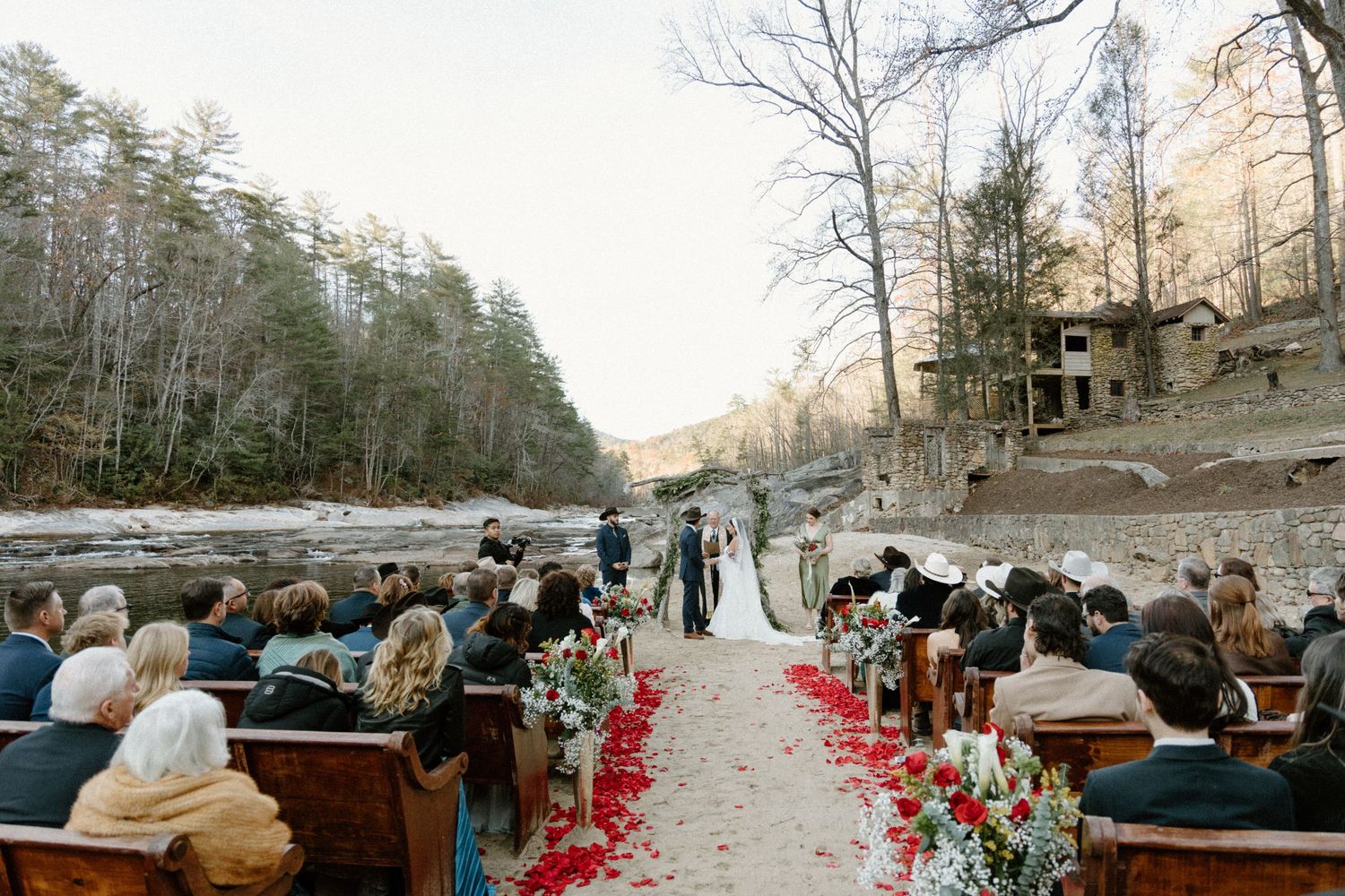 A winter forest ceremony alongside a river with wooden benches and red rose petals lining a pathway in the woods.