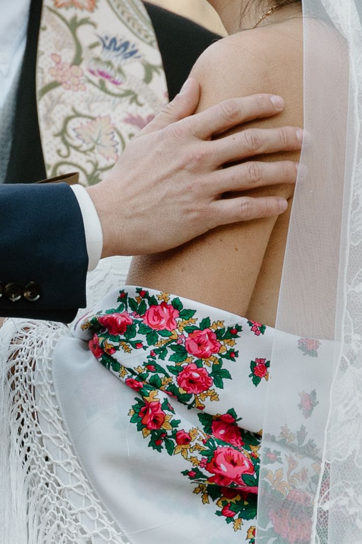 Close-up detail of a wedding dress with embroidered red roses and lace trim as a groom's hand rests on the bride's arm.