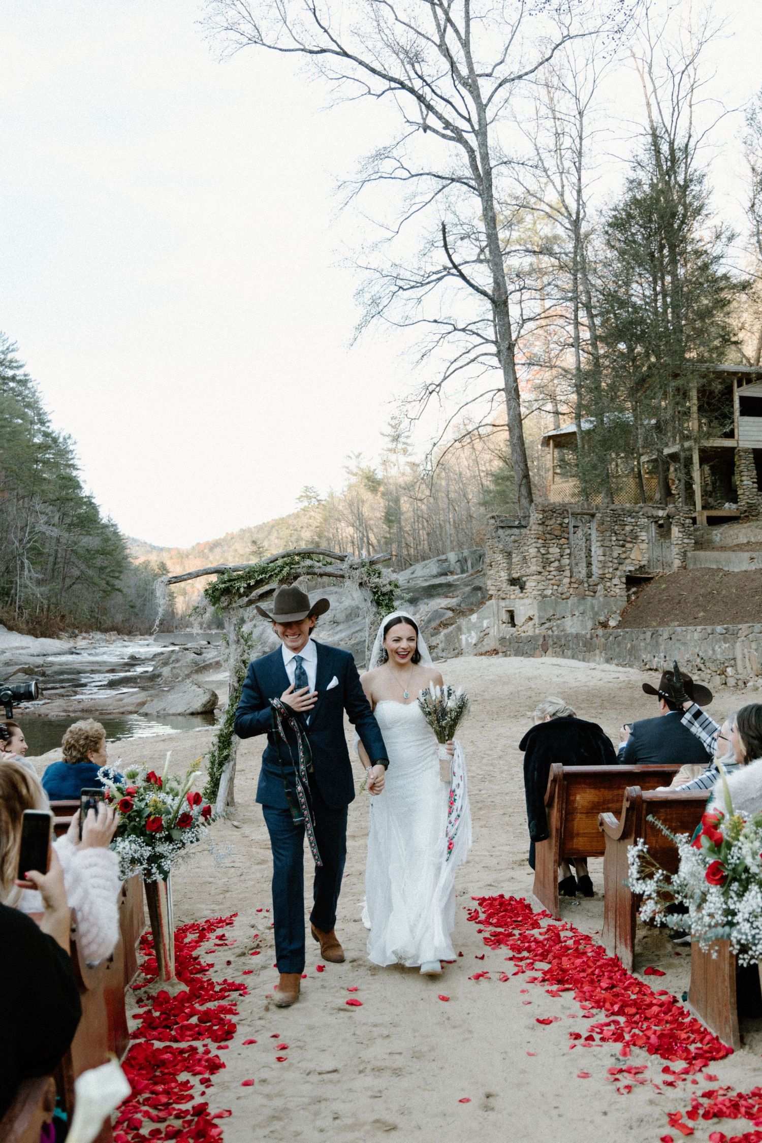 A winter wedding ceremony with scattered red rose petals along the aisle in a natural outdoor setting.