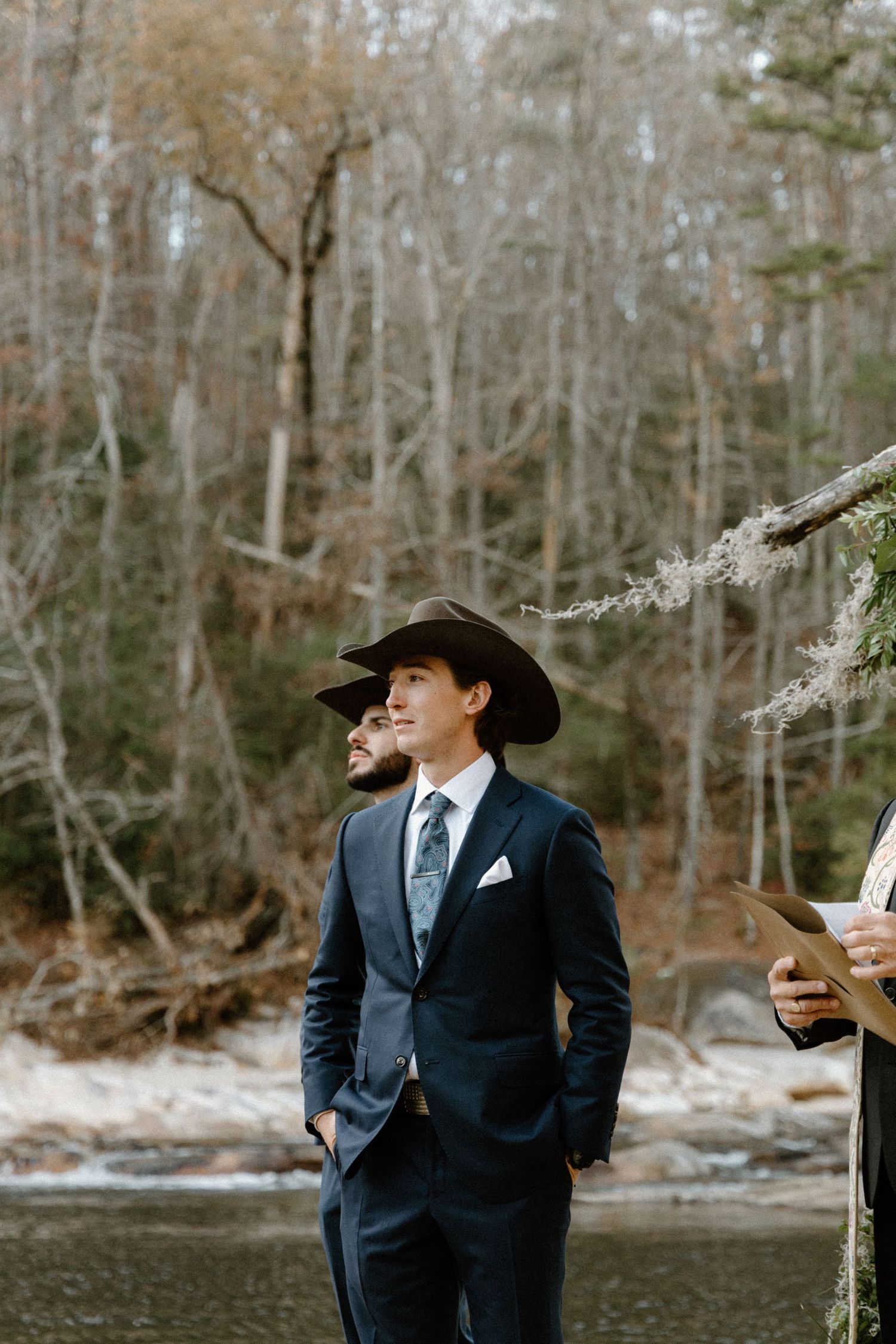 A figure in a dark suit and cowboy hat stands by a calm lake surrounded by winter woods on a peaceful afternoon.