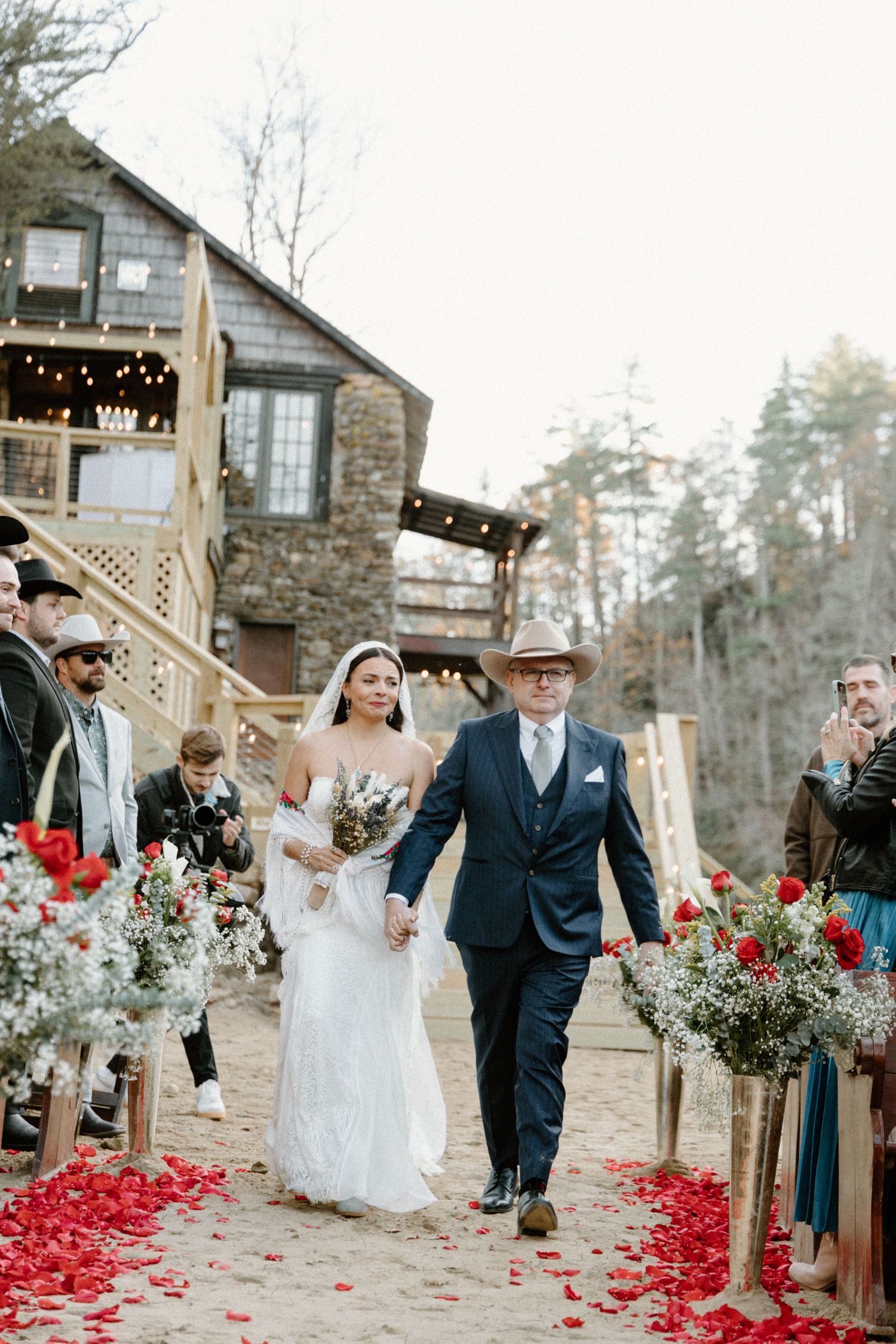 Outdoor wedding ceremony with red rose petals scattered along the aisle and rustic stone building in the background.