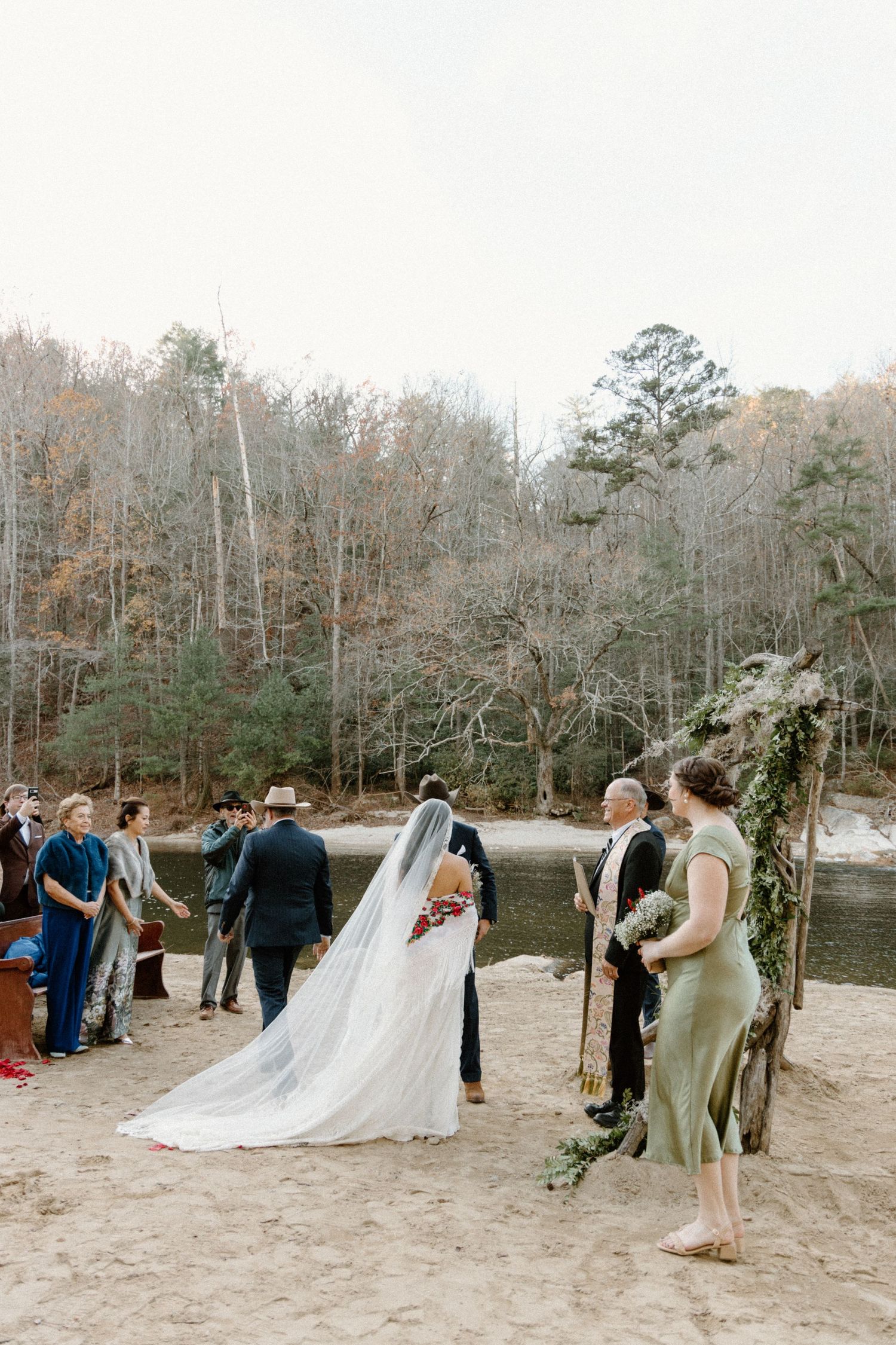 A wedding ceremony takes place on a sandy beach with guests gathered beside bare winter trees and a tranquil lake.