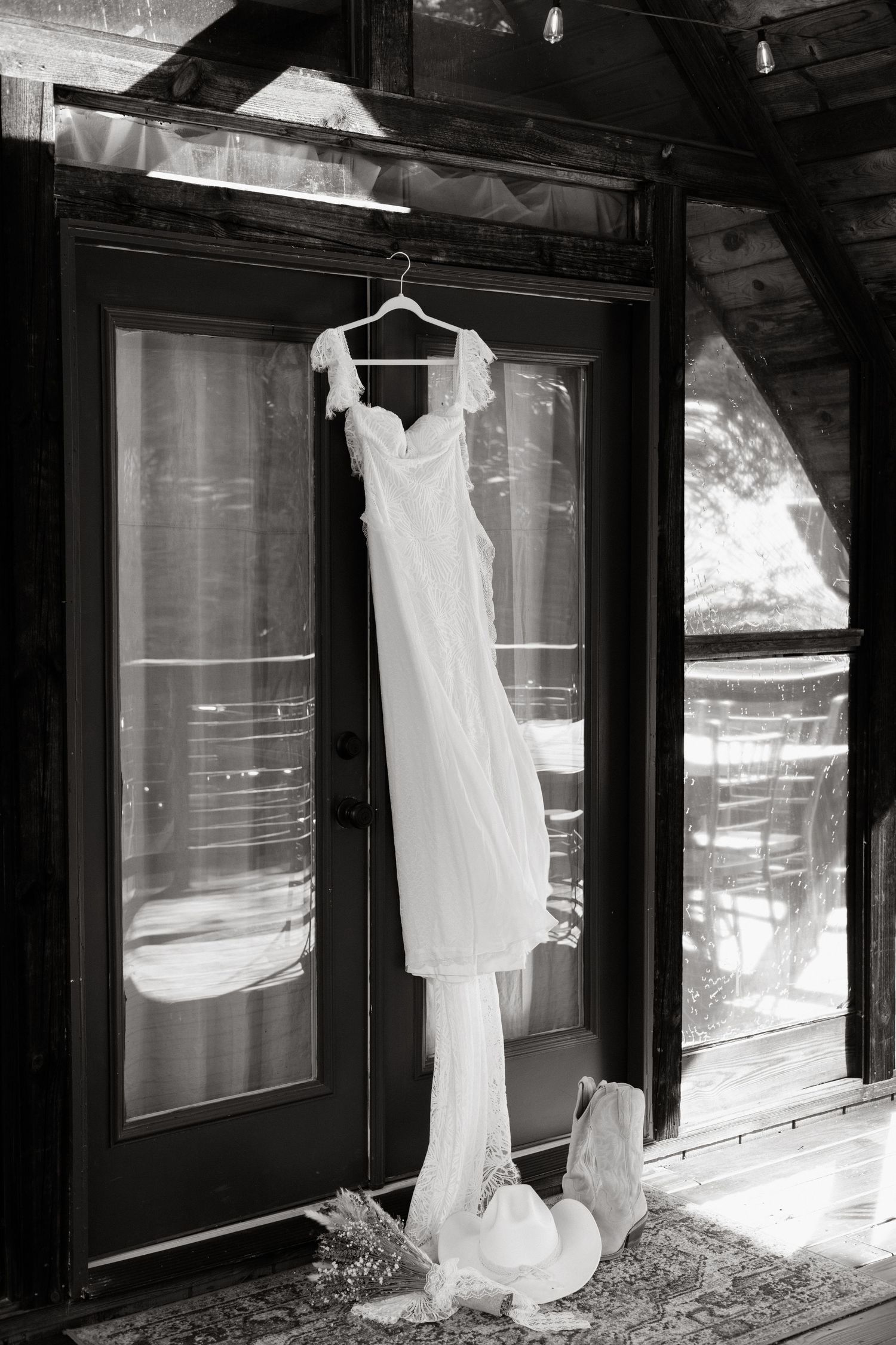 An elegant white wedding dress hangs on a wooden hanger against glass doors in black and white photography.