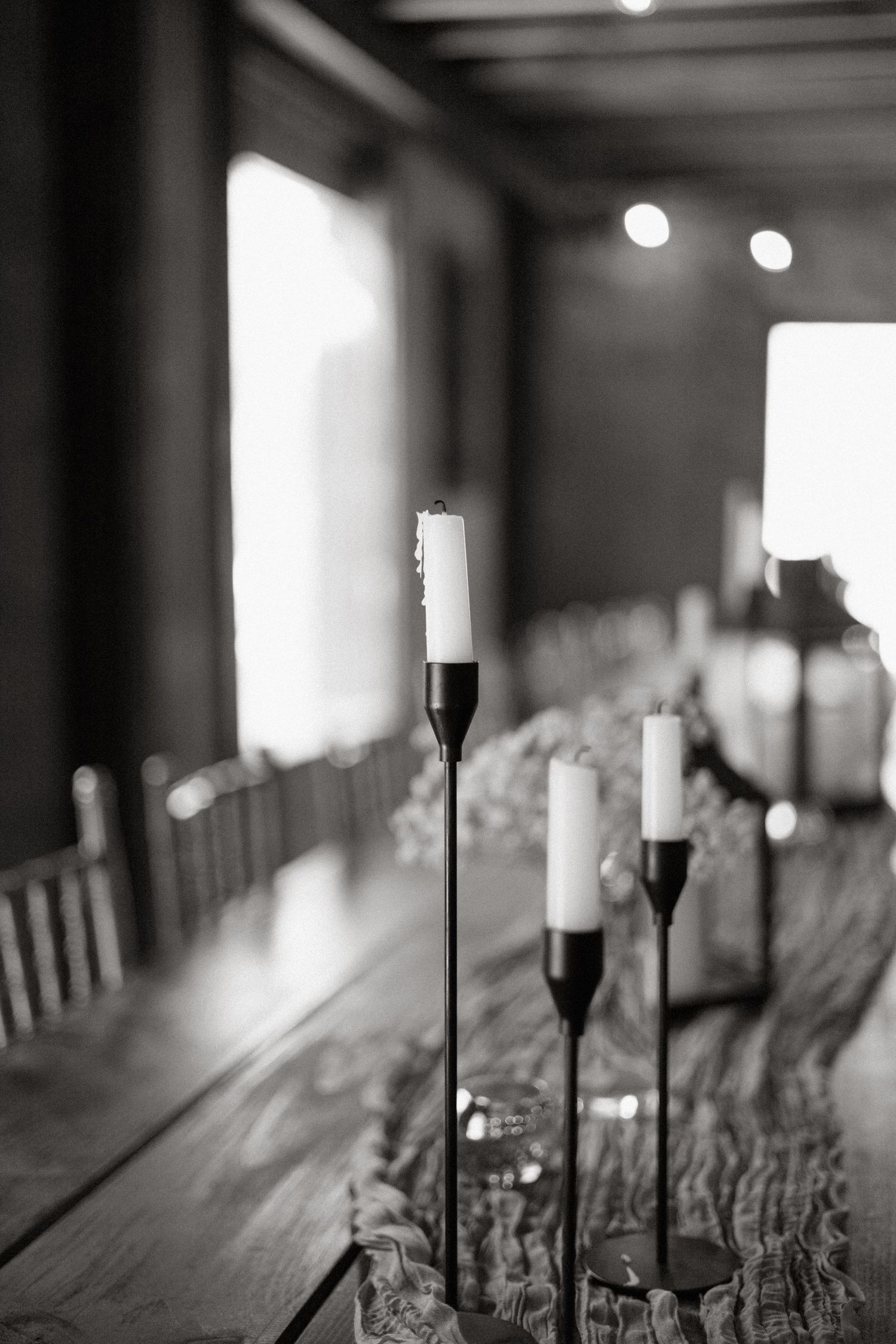 Black and white photo of tall candlesticks along a rustic wooden table with bokeh lights in the background.