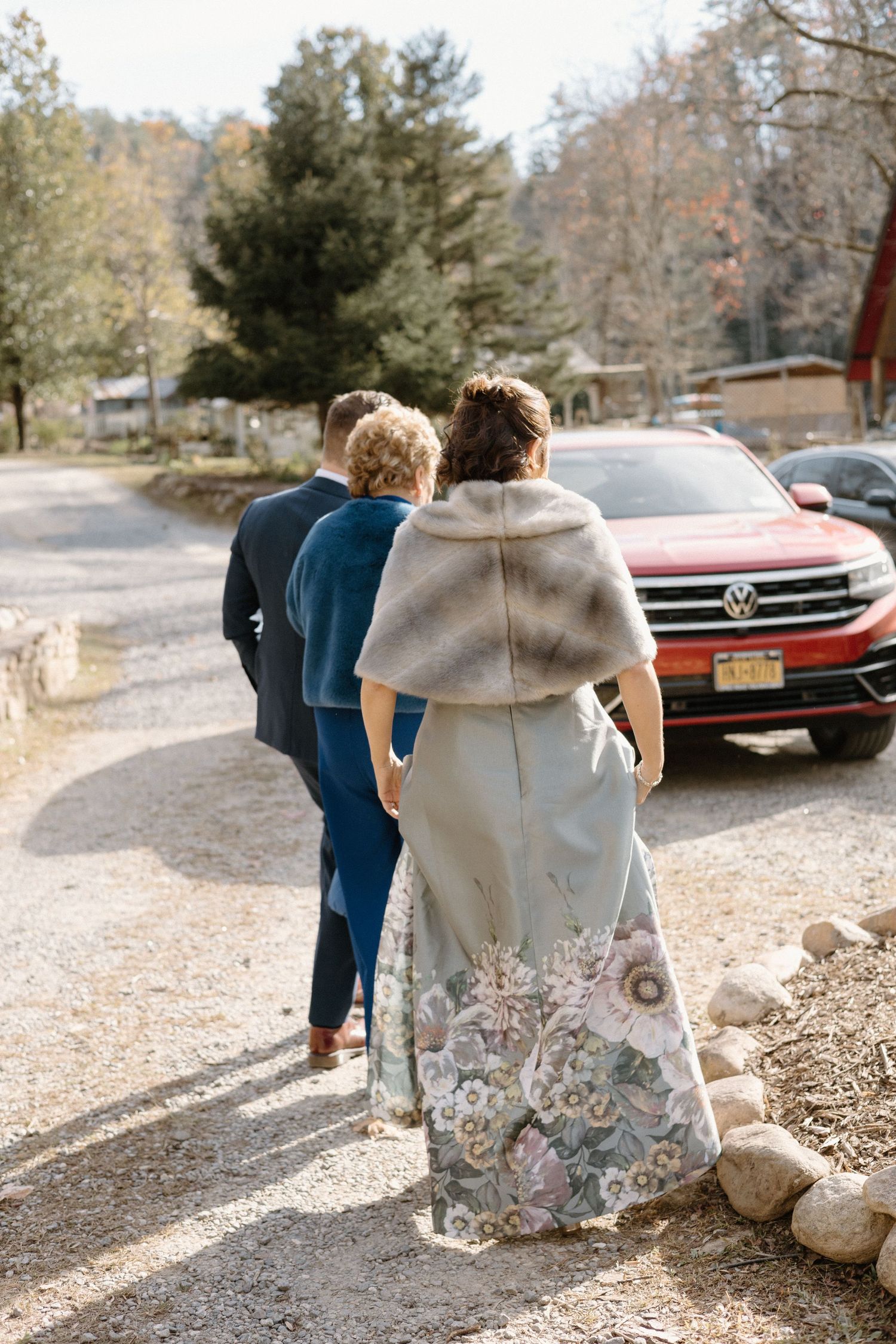Three people walking on a gravel path next to a red car with autumn trees and a rural setting in the background.