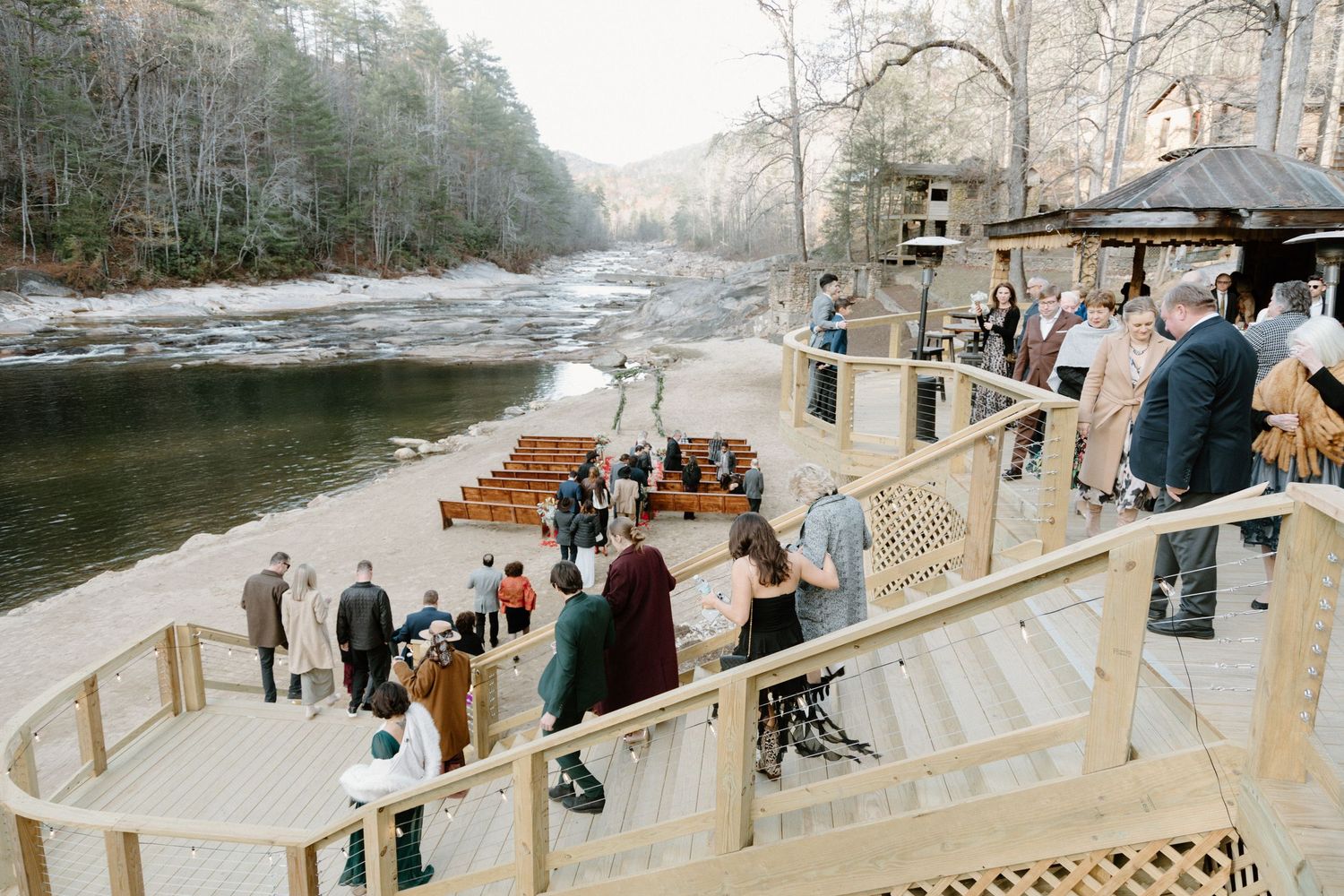Wedding guests gather on a curved wooden deck overlooking a misty winter river in a serene forest setting.