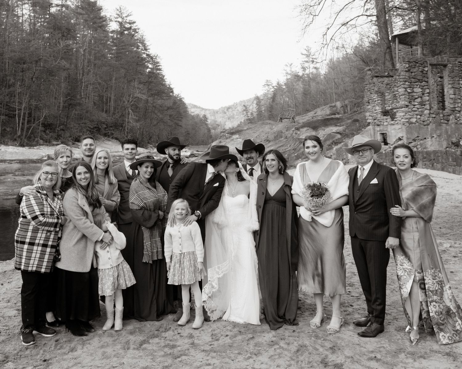 Group of people gathered outdoors at mountain wedding celebration with rustic stone wall and forest backdrop.