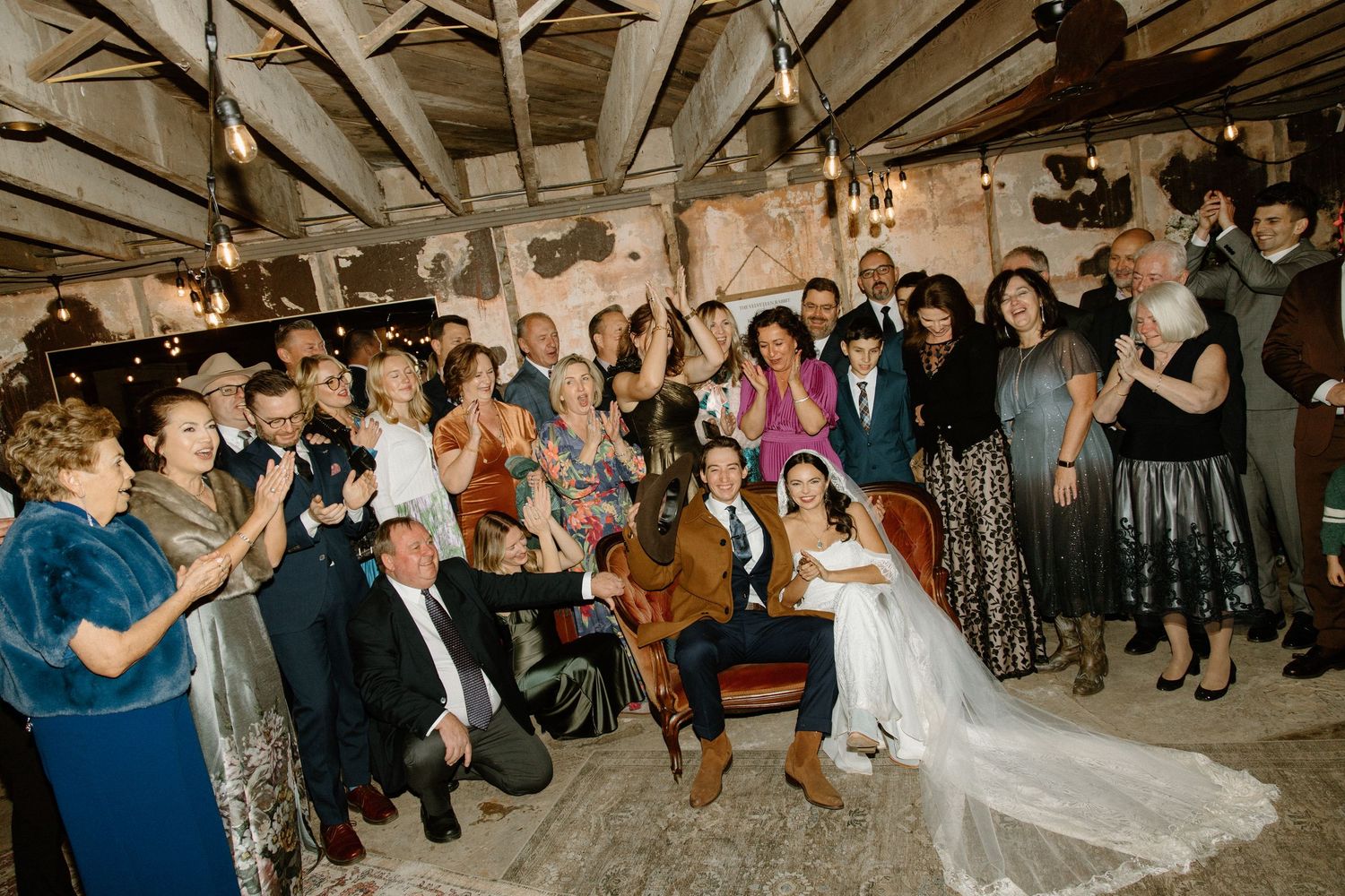 Wedding guests clap and cheer during a celebration in a rustic barn venue with exposed wooden beams and warm lighting.