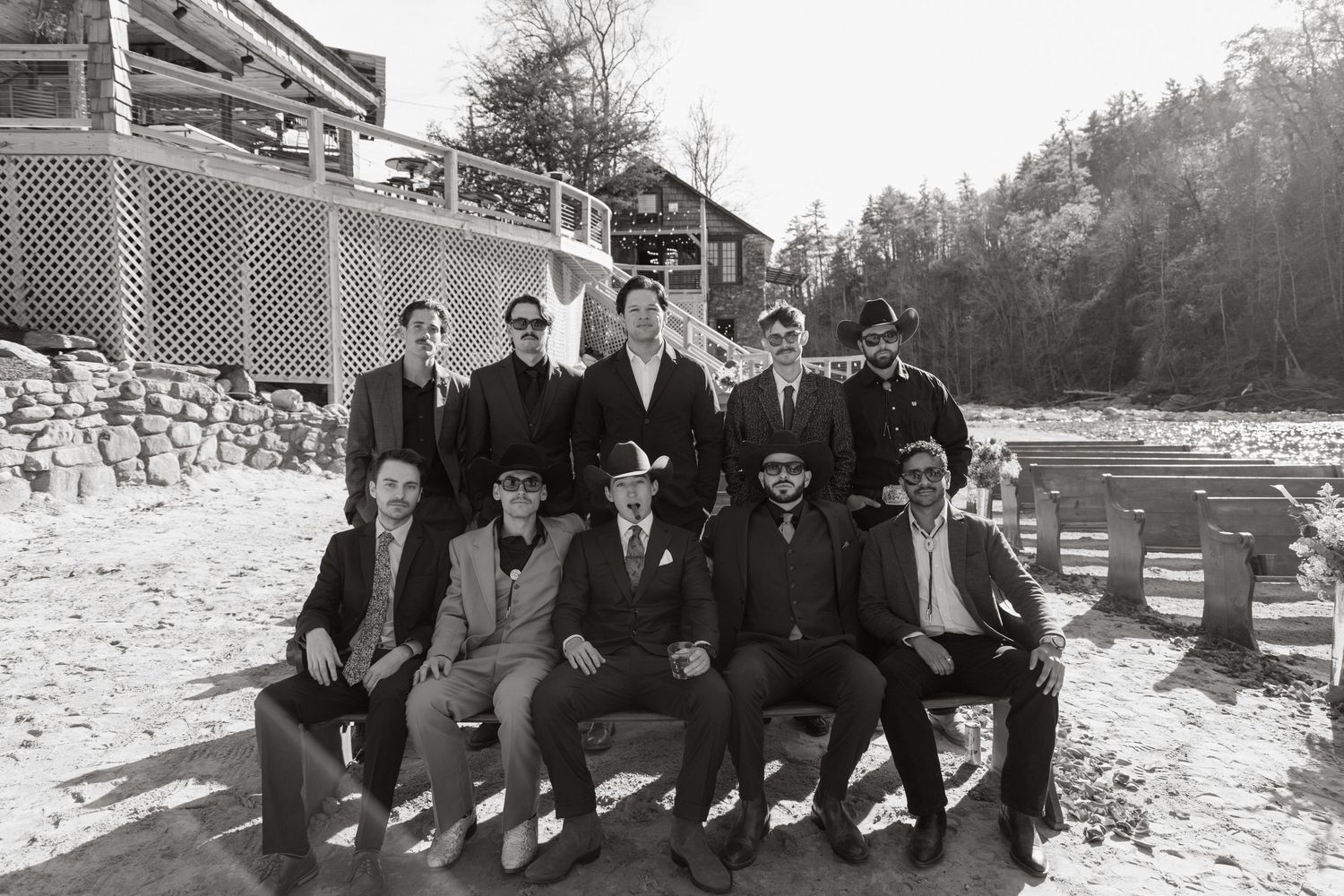 A group of men in formal attire seated on a bench against a snowy backdrop near a stone wall and wooden building.