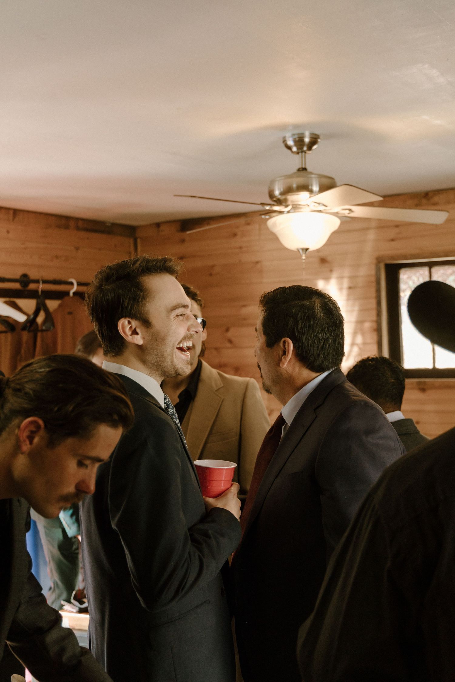 People gathered at an indoor social event sharing laughs and conversation in a wood-paneled room with ceiling fan lighting.