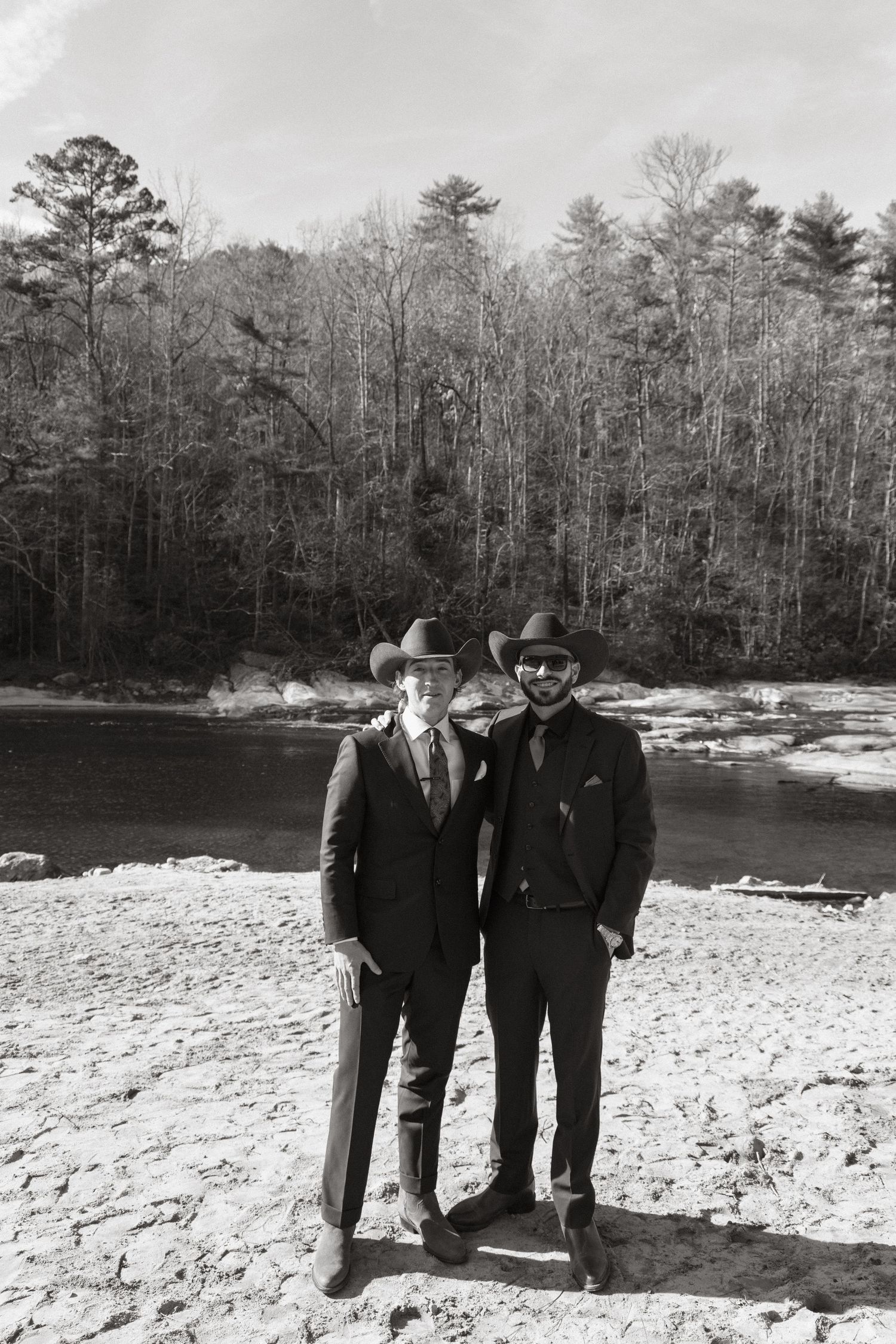 Black and white photograph showing two figures in cowboy hats standing on a rocky riverbank surrounded by winter trees.