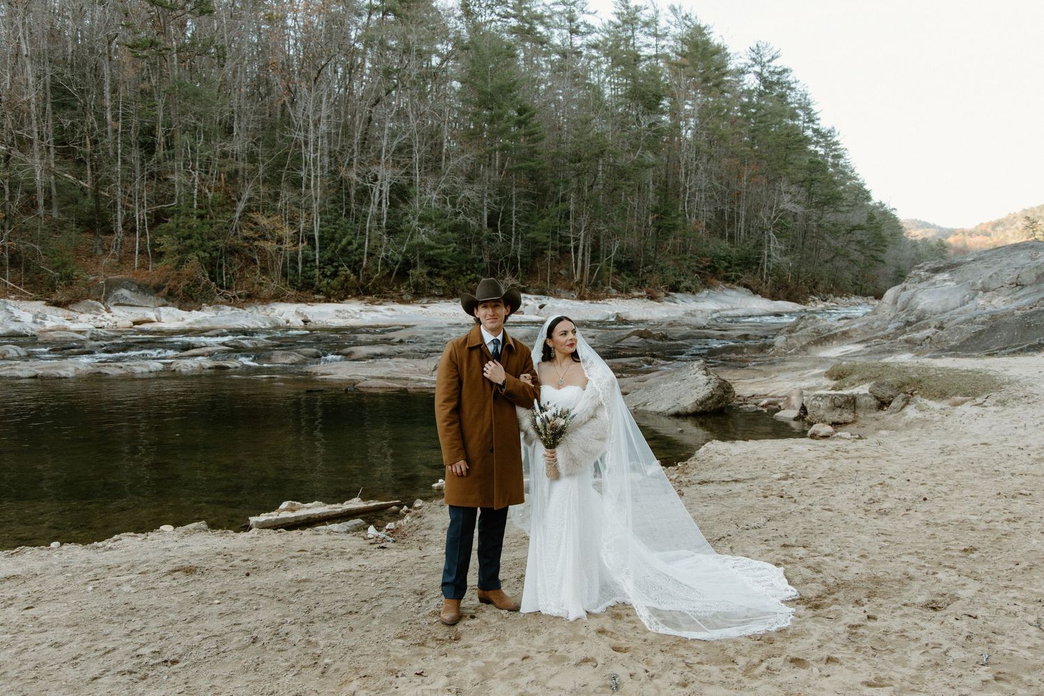 A couple poses together on a rocky riverbank with pine forest and water in the background on a winter day.