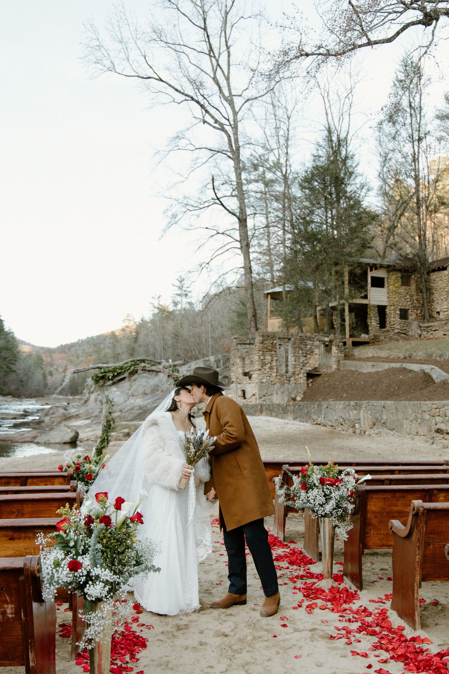 Couple embraces at rustic outdoor wedding ceremony with wooden benches and red rose petals scattered on the ground.