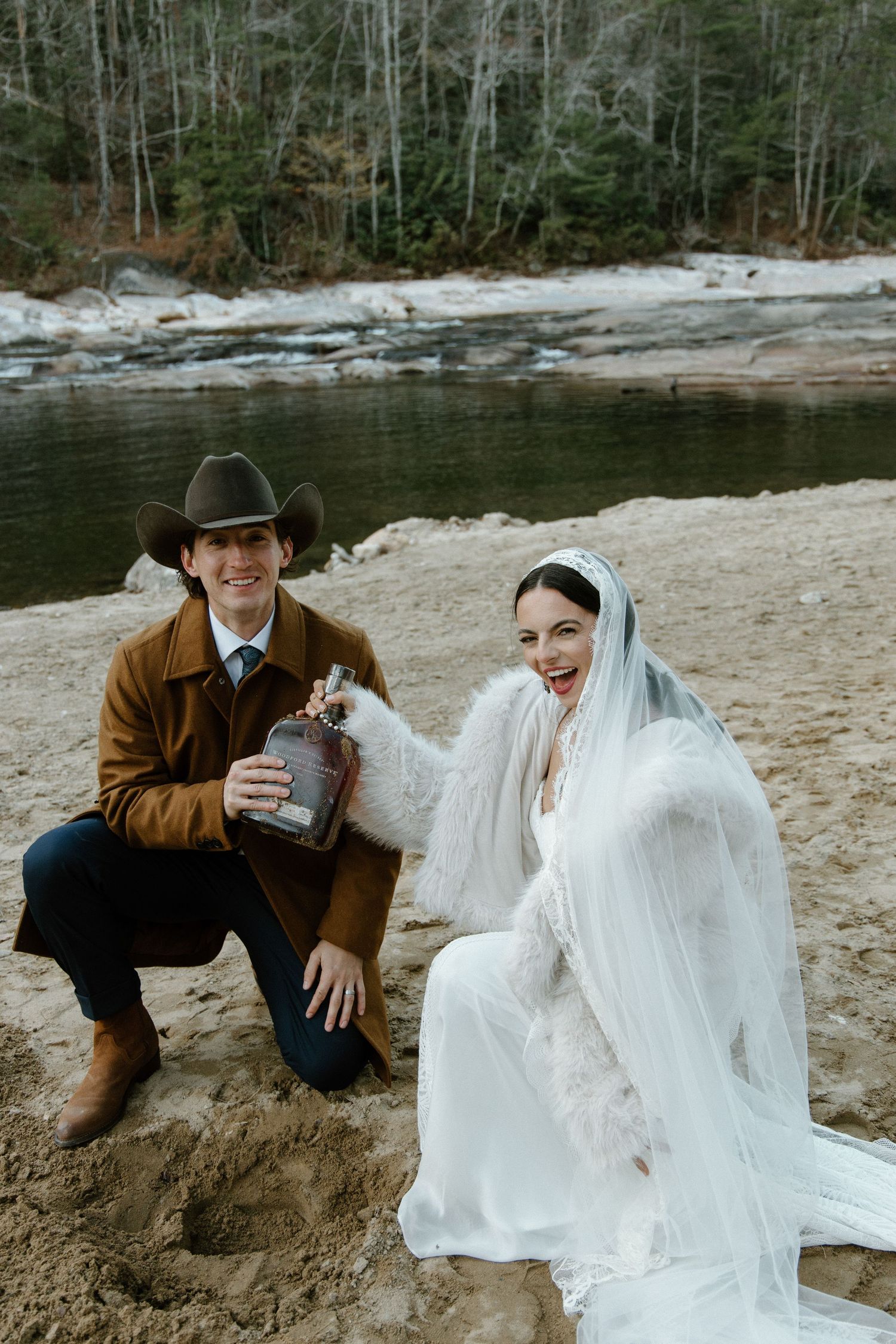 A couple in wedding attire shares intimate moments by the water's edge on a sandy beach surrounded by trees.