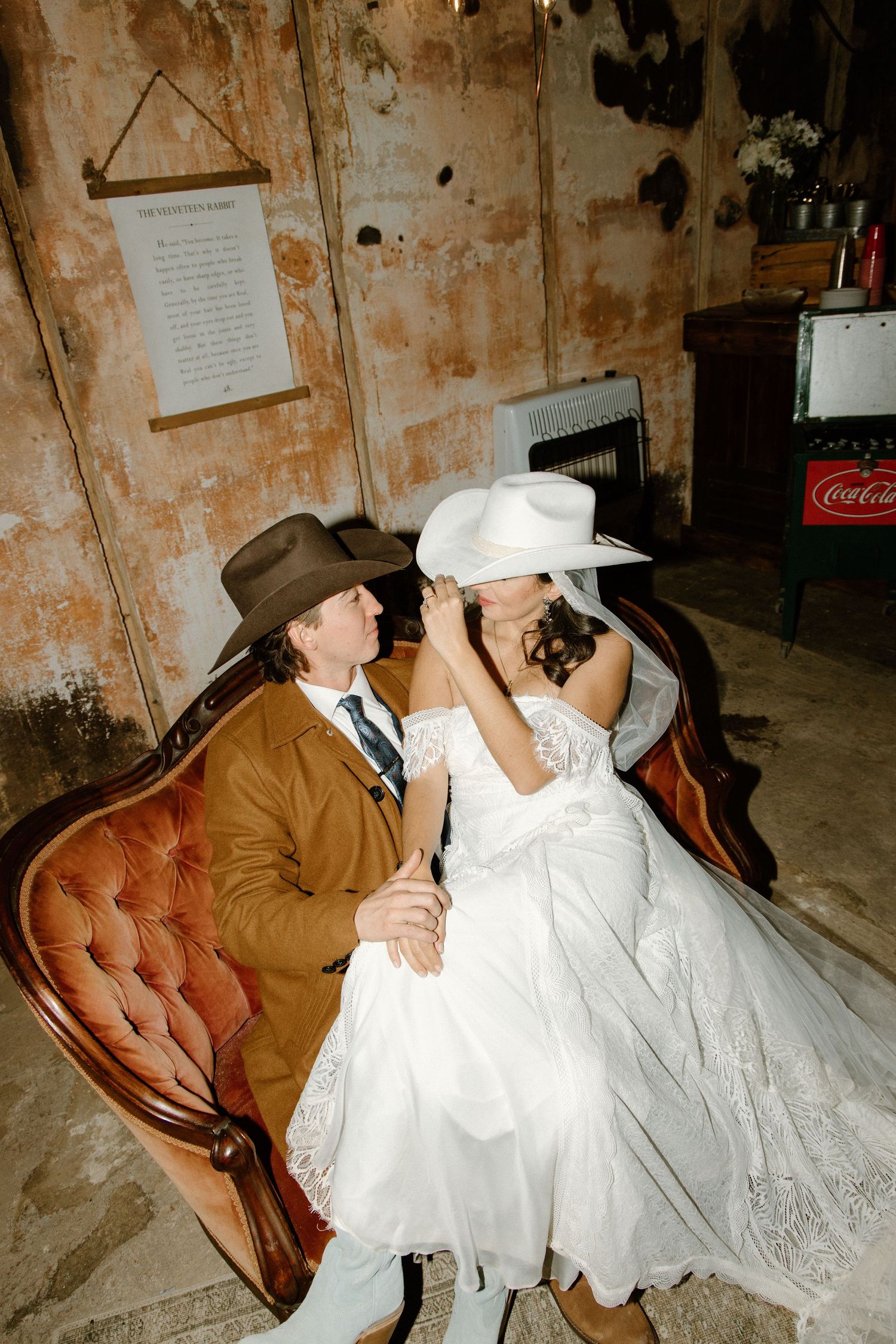 A couple in western attire sharing a vintage leather couch, with the bride in a white dress and both wearing cowboy hats.