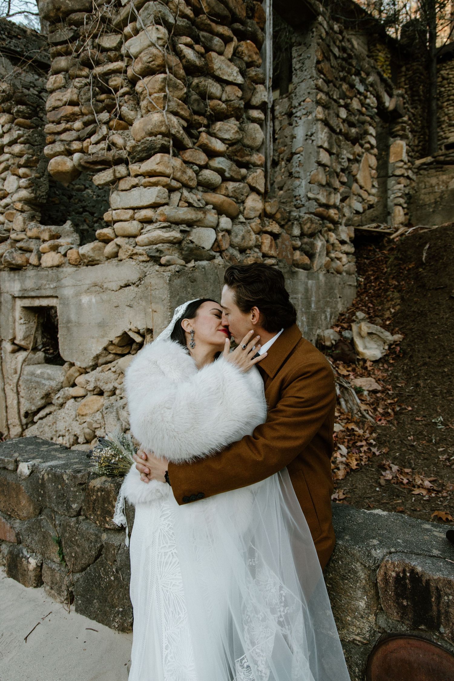 A couple embraces against a rustic stone wall backdrop on their wedding day, with bride wearing white dress and fur shawl.