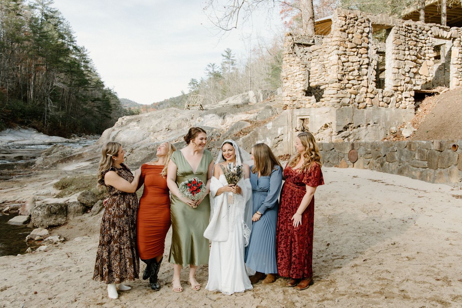 Six people in colorful dresses stand together against a rocky cliff wall outdoors near a forest landscape.